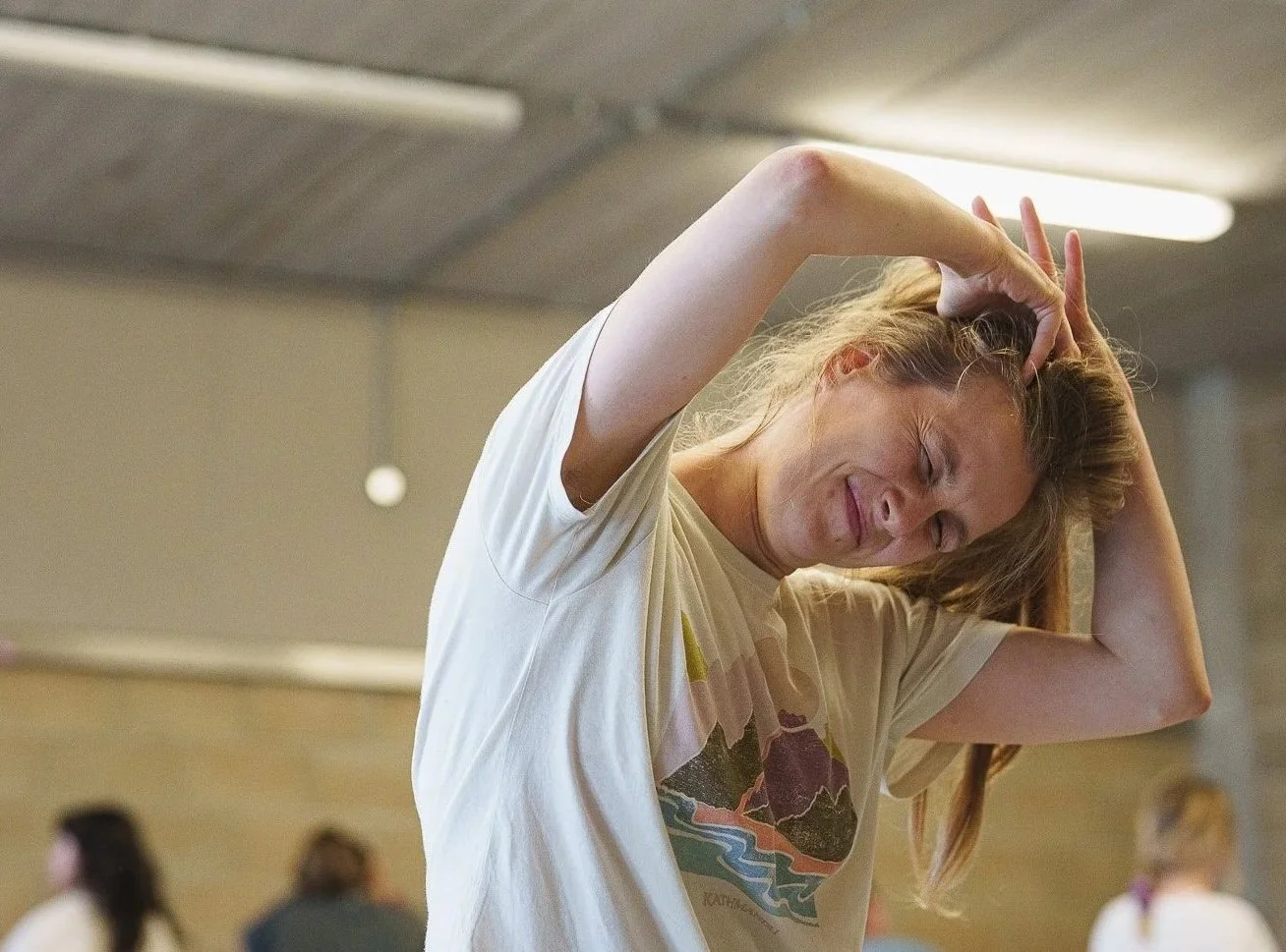A woman with blonde hair in a beige t-shirt is stretching with her hands on her head and eyes closed, in an indoor space with others in the background.