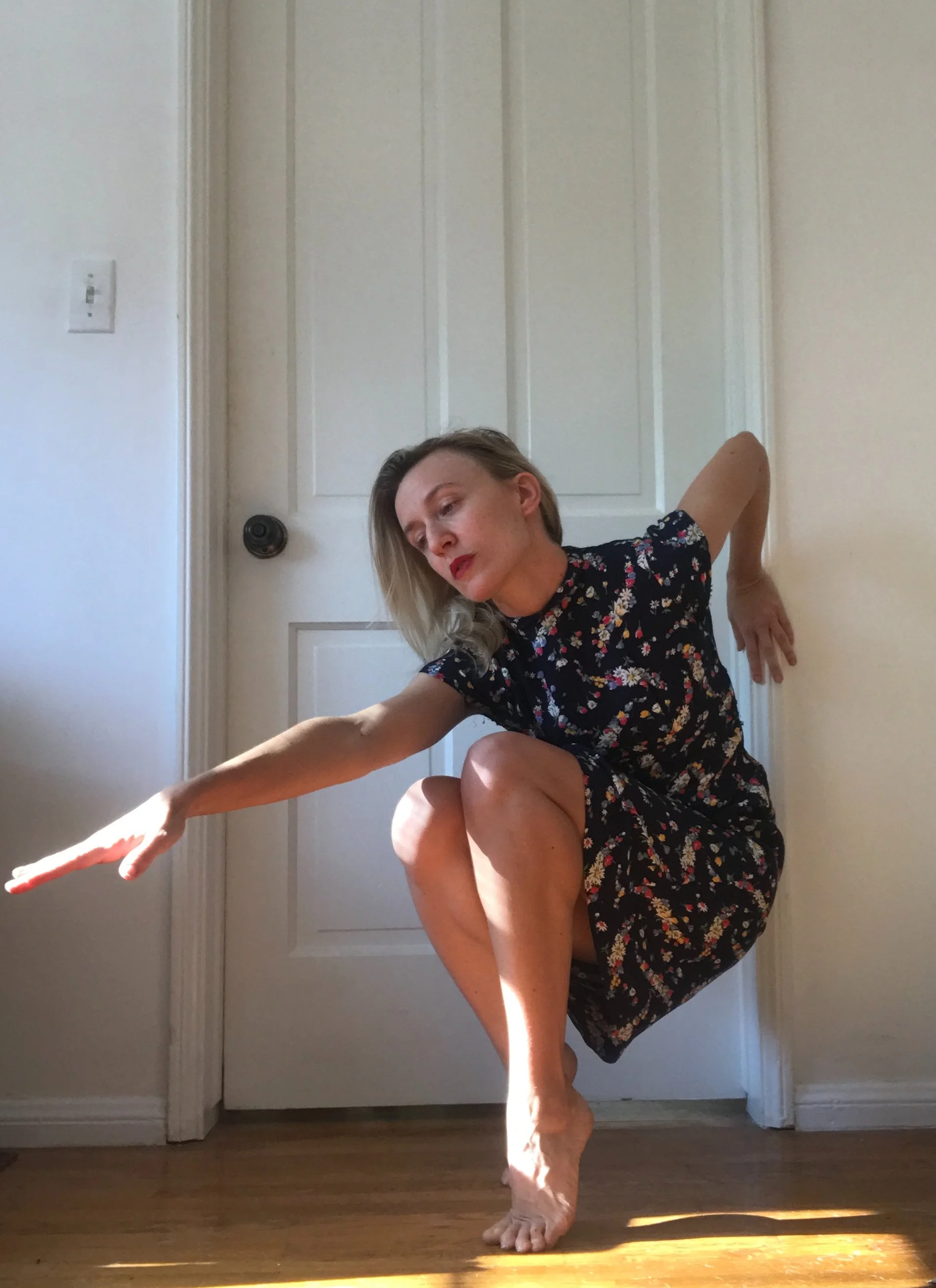 A woman with blonde hair, wearing a floral dress, is practicing a dance pose indoors near a closed door, balancing on one foot with her other leg bent and arms extended gracefully.