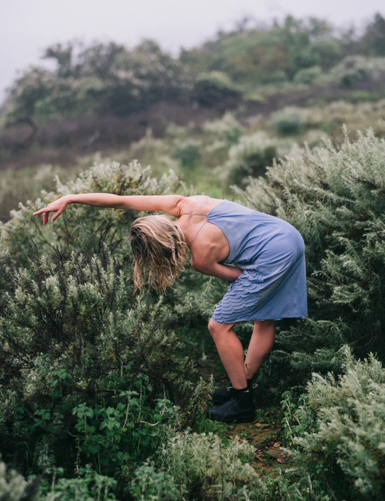 Woman in a blue dress practicing yoga outdoors among lush green bushes.