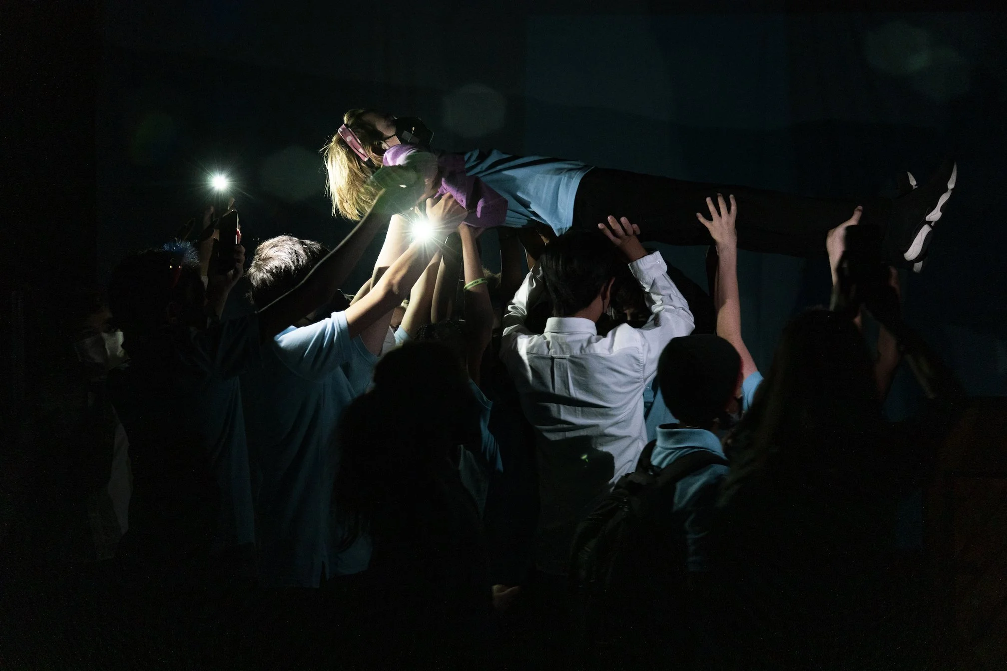 Group of students lifting a girl in a blue hoodie while taking photos in a dark room illuminated by camera flashes.