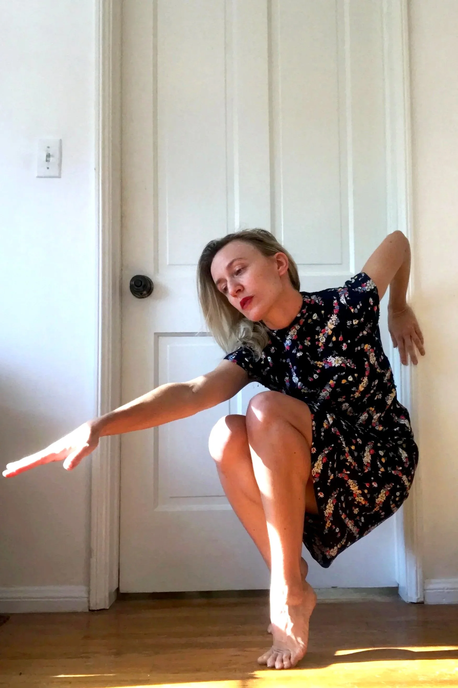 Woman in a floral dress balancing on one foot with her arms extended, indoors near a white door and sunlight on the wooden floor.