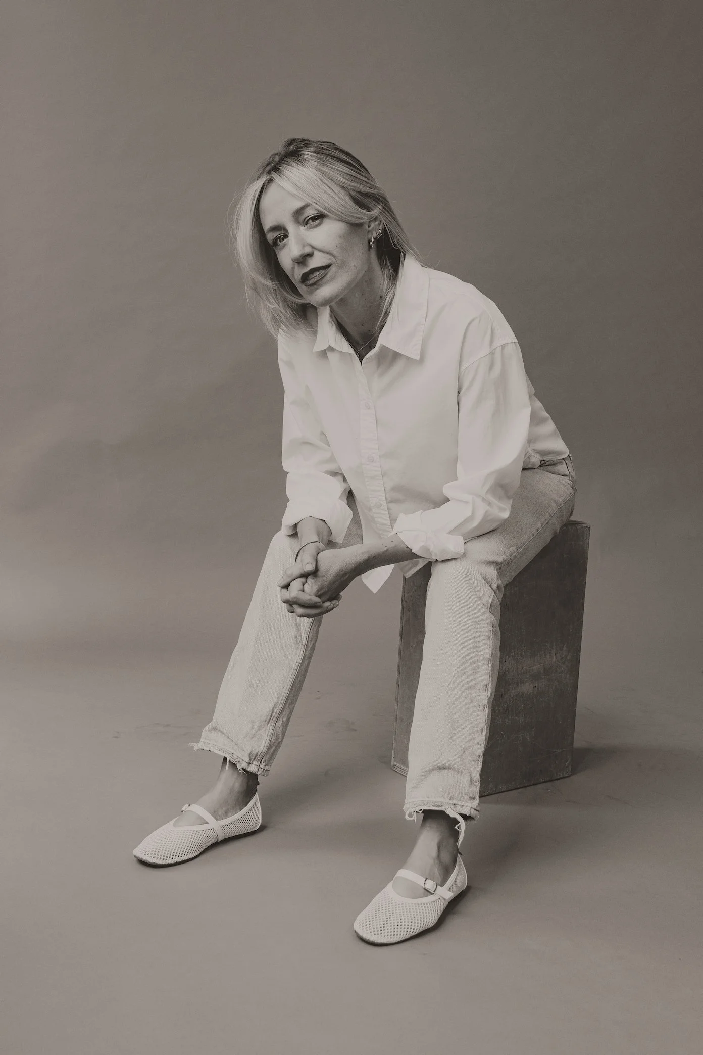 A black-and-white photo of a woman sitting on a wooden block against a plain background. She is wearing a white shirt, light-colored jeans, and white shoes. She has blond hair, some jewelry, and is looking at the camera with a slight smile.