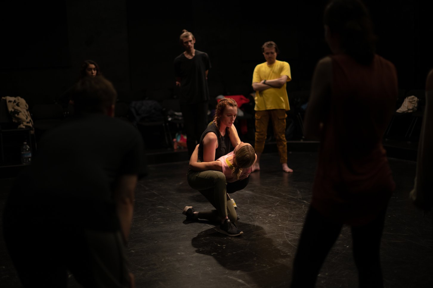 People rehearsing or performing on a dark stage, with one person kneeling and holding another person, surrounded by others standing and observing.