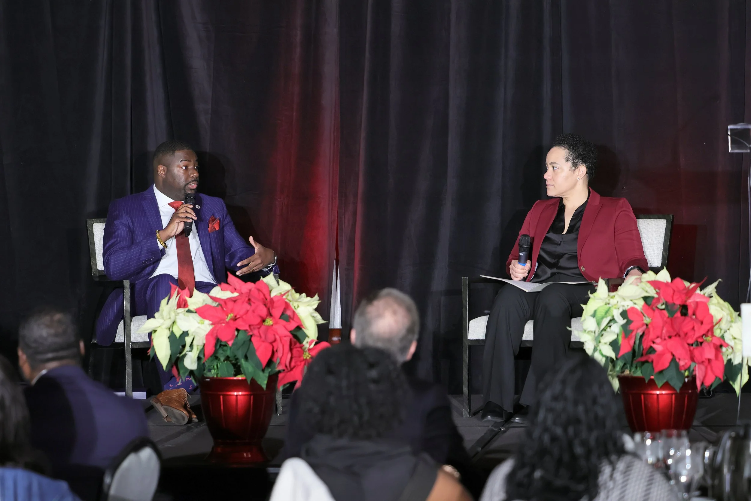 A panel discussion with two speakers on stage, one man and one woman, each holding a microphone, seated in front of black curtains and decorated with poinsettia plants, with an audience watching.