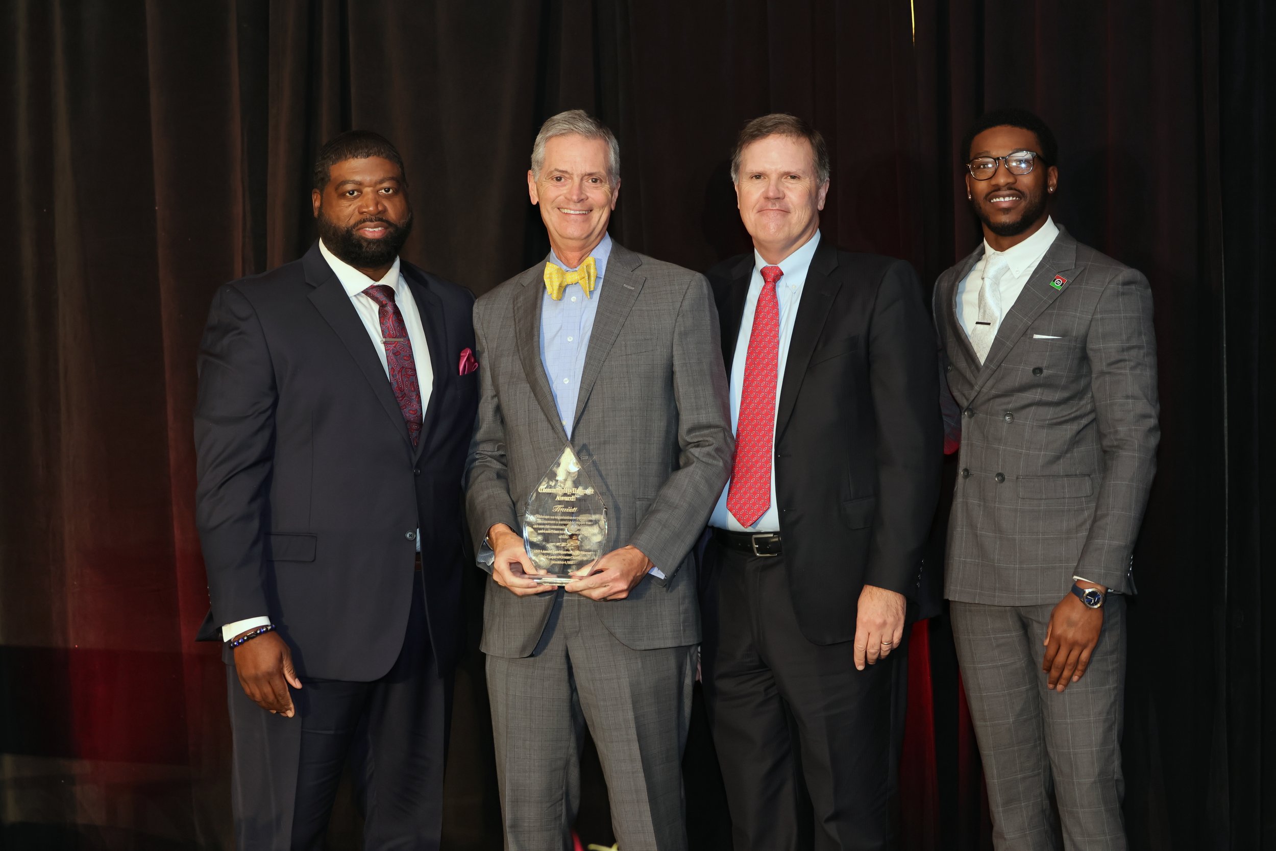 Group of four men in suits standing together on stage, with one holding an award, at a formal event or ceremony.