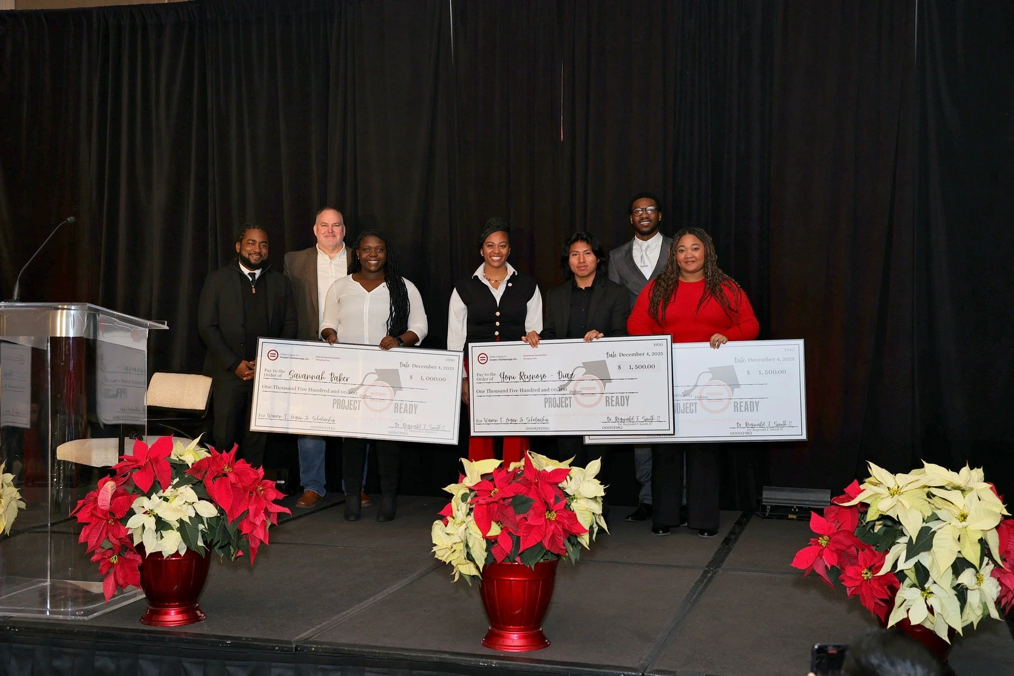 Group of seven people standing on stage holding oversized checks, with festive red and white poinsettias in front, black curtains in the background, and a podium on the left.