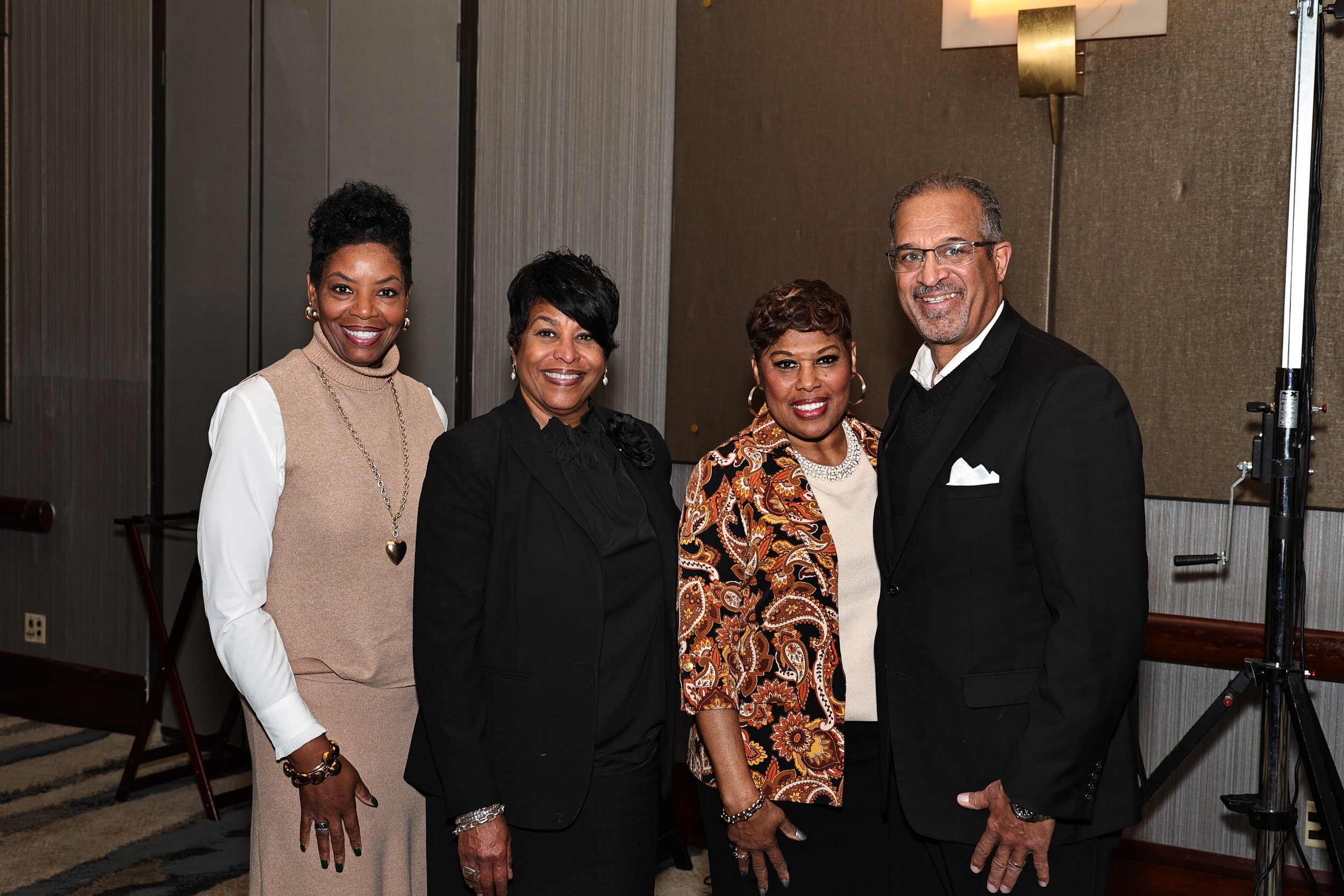 Four diverse adults dressed in business or semi-formal attire smiling and posing together at an indoor event.