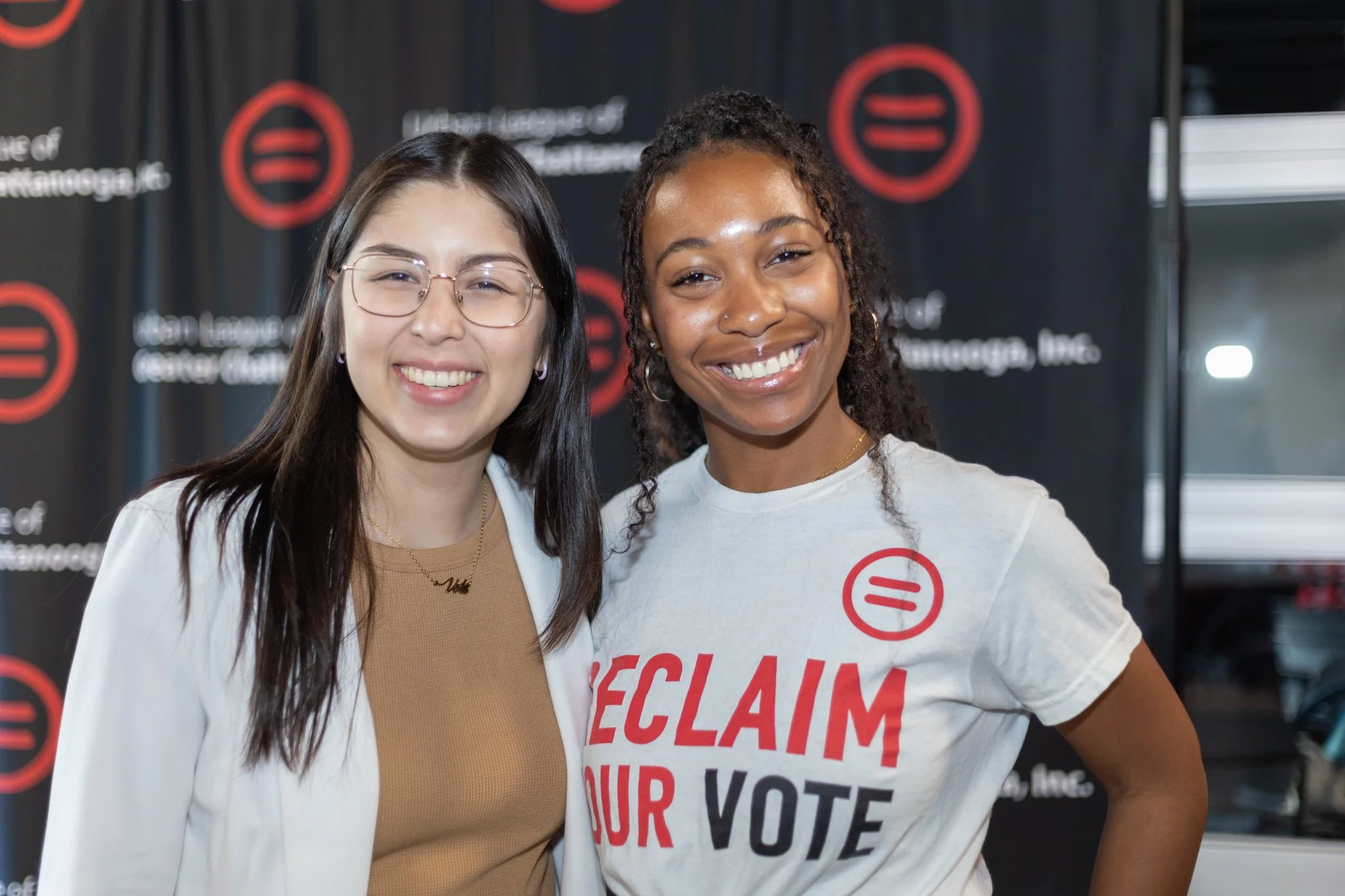 Two smiling women standing together, one wearing glasses and a white blazer, the other wearing a white t-shirt with red and black text that says 'RECLAIM YOUR VOTE,' posing in front of a black backdrop with red and white logos and text.