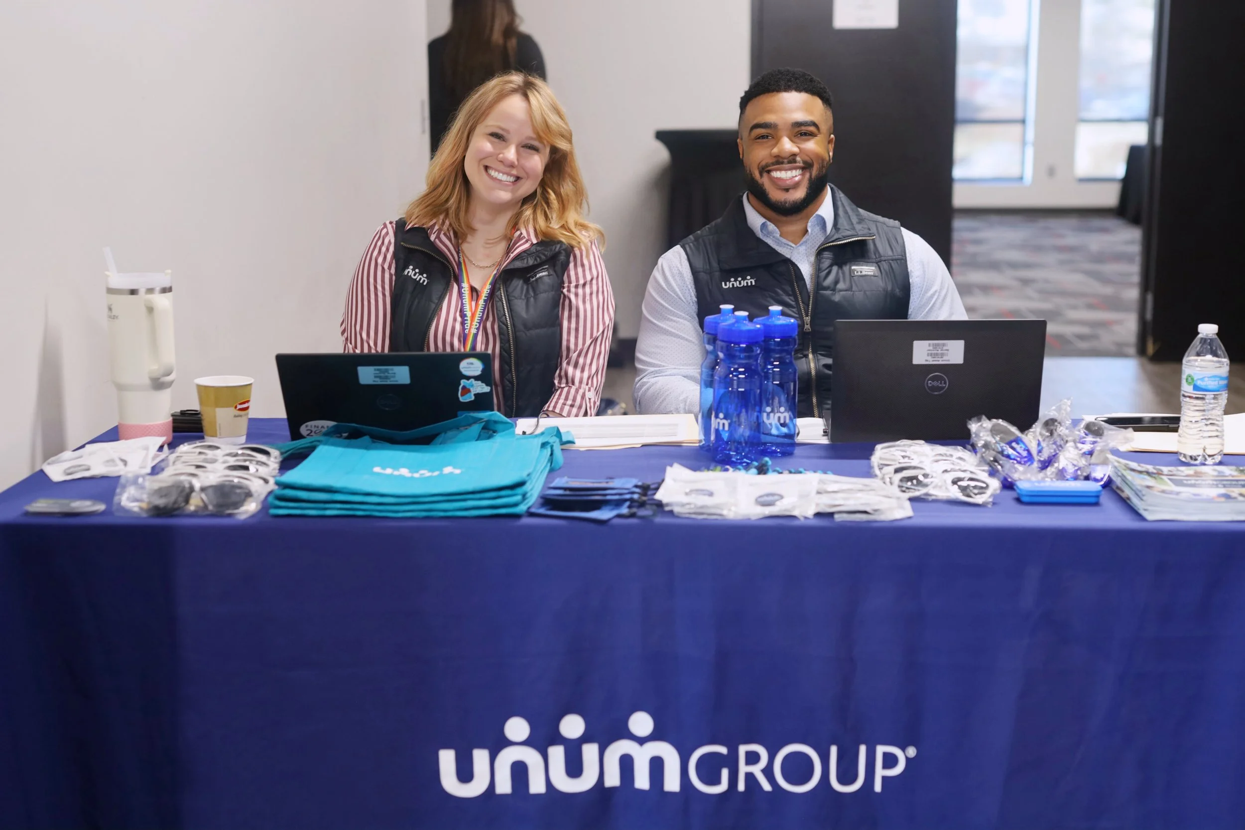 Two smiling people sitting at a registration table with promotional materials, water bottles, and laptops, branded with Unum Group, in a conference or event setting.