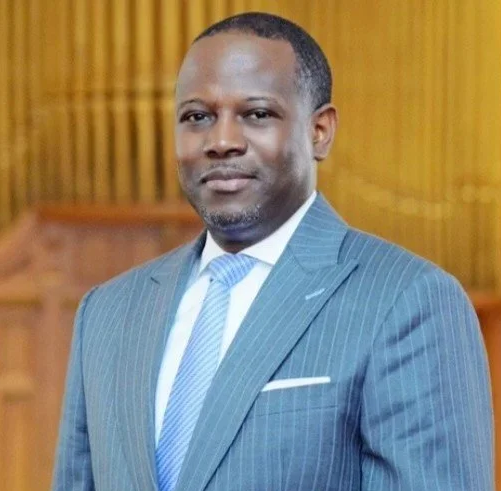 A man in a blue pinstripe suit with a light blue tie standing in an indoor setting with wooden paneling in the background.