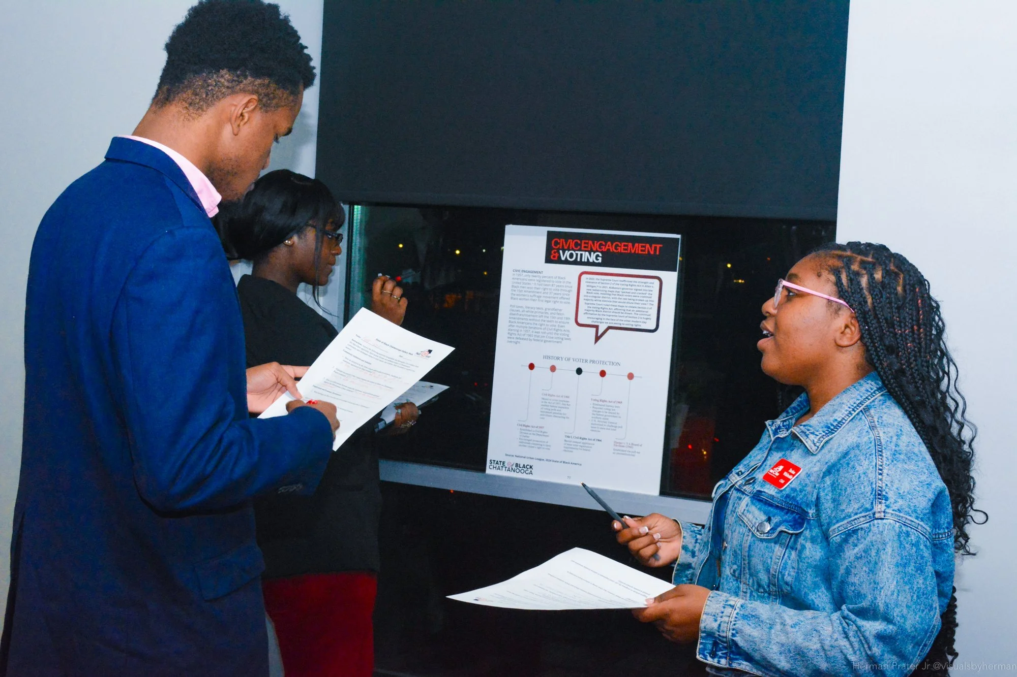 Three people standing near a display poster about civic engagement and voting, engaging in discussion and reading documents.