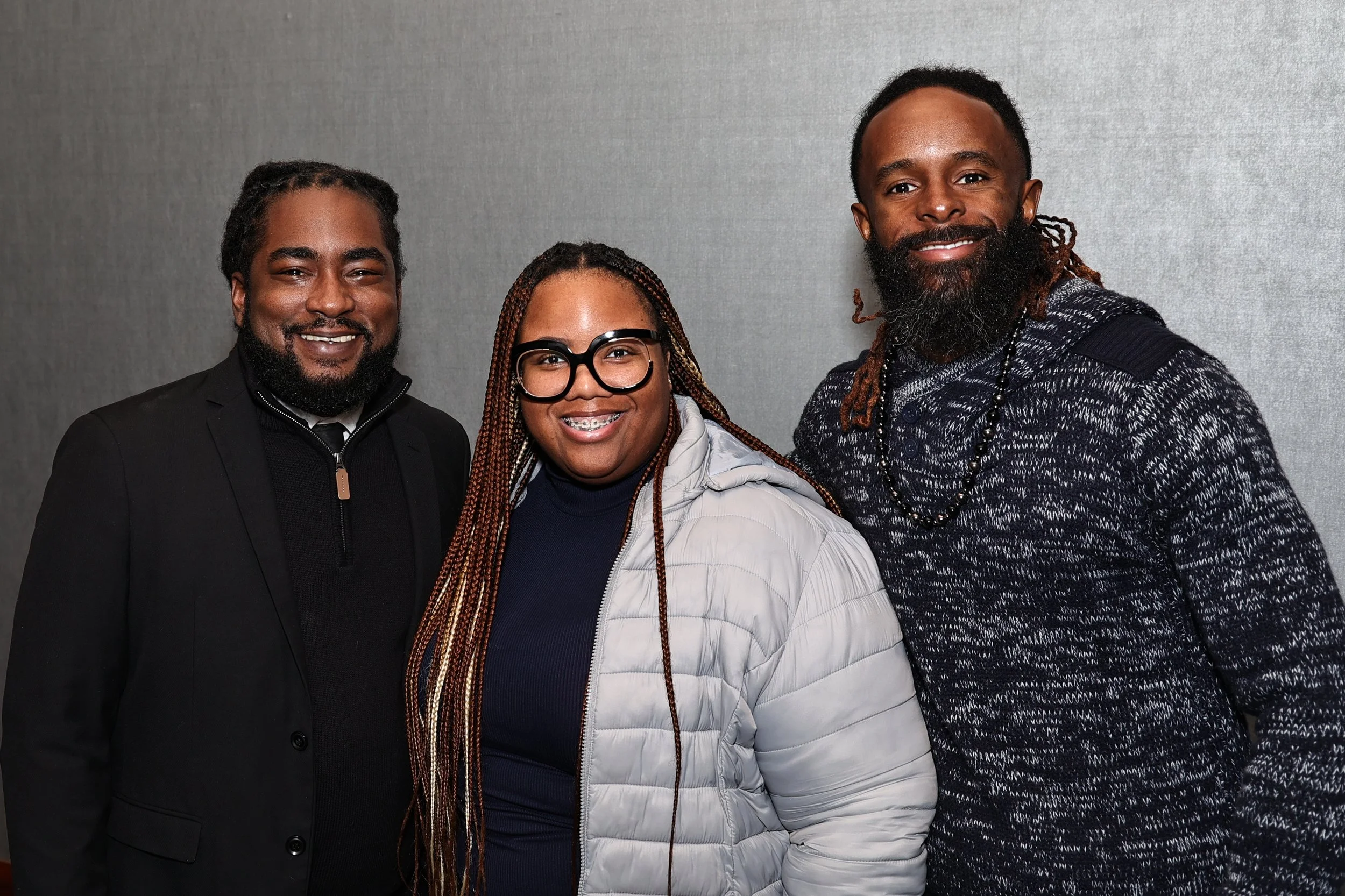 Three people standing together indoors in front of a gray textured wall, smiling at the camera.