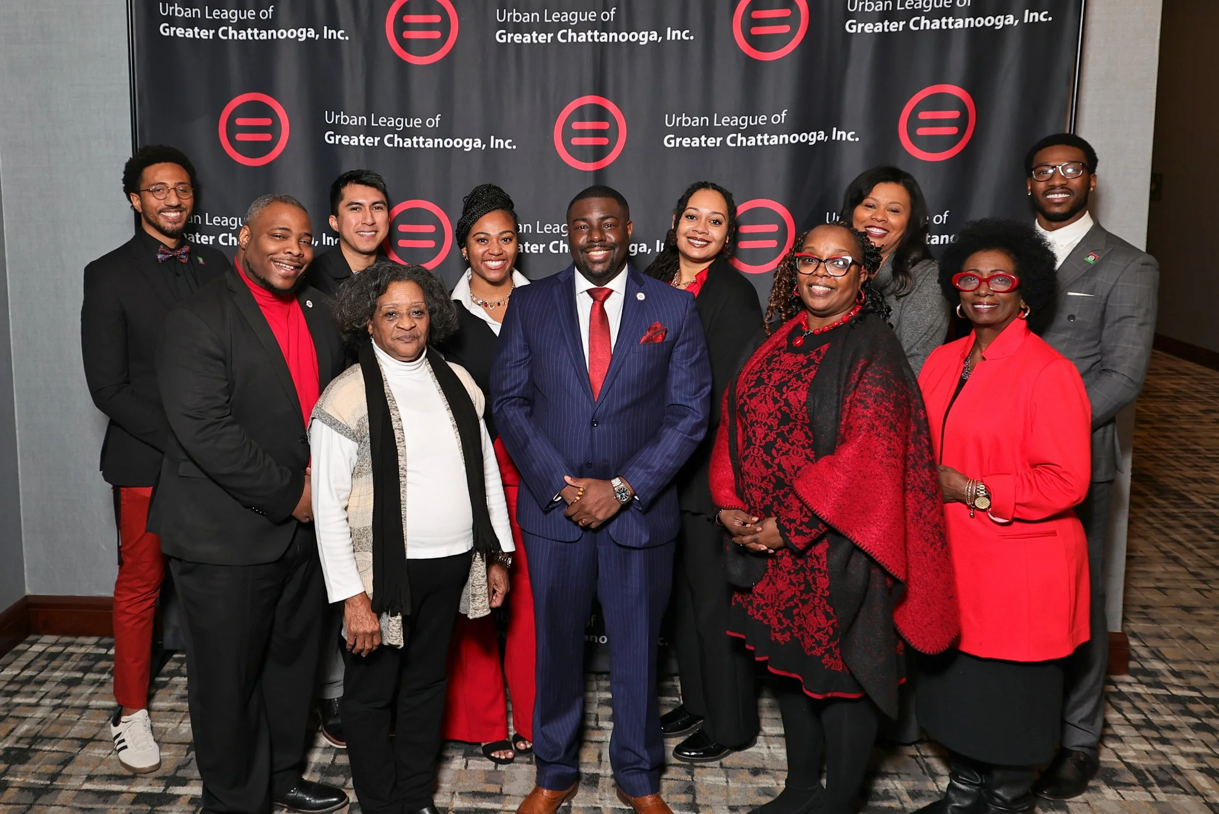 Group of diverse professionally dressed people posing in front of a black backdrop with red and white logo and text for Urban League of Greater Chattanooga, Inc.