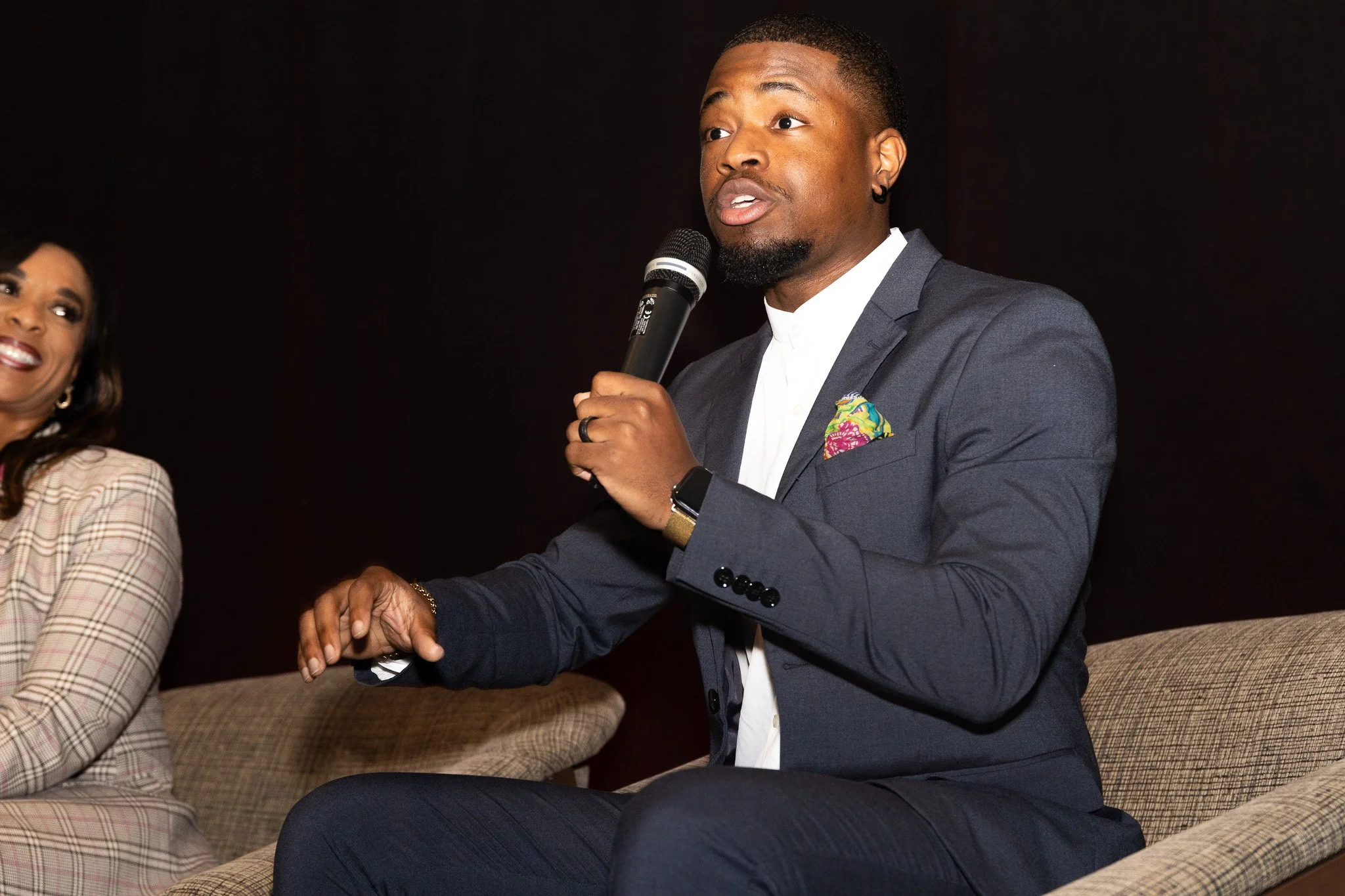 A man in a dark suit and white shirt holding a microphone speaks while seated on a beige upholstered chair, with a woman smiling beside him against a dark background.