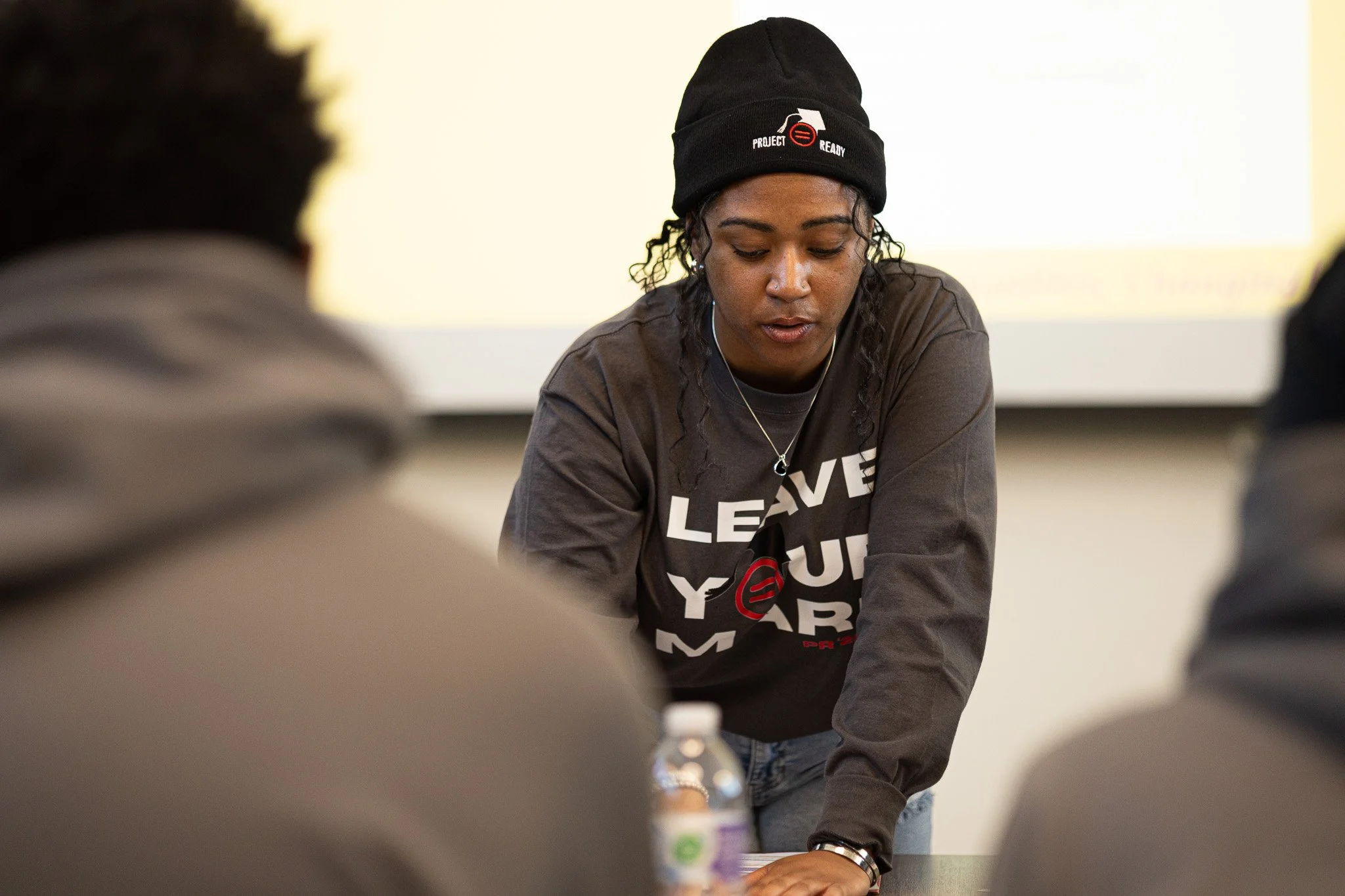 A young woman wearing a black beanie and a gray shirt leaning over a table during a discussion, with a water bottle and two people blurred in the foreground.