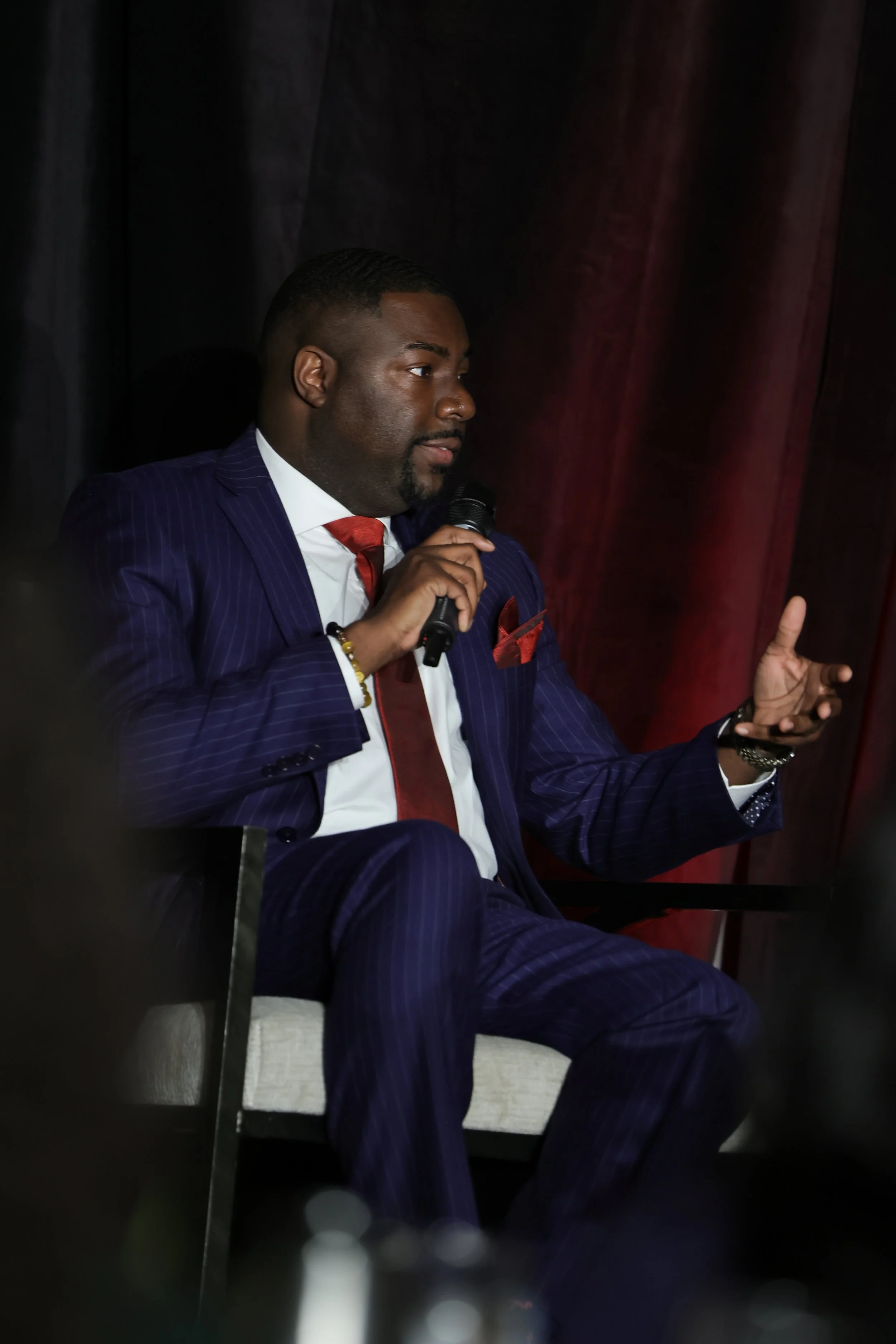 A man in a dark blue pinstripe suit, white shirt, and red tie sitting with a microphone in his hand, gesturing with his other hand, in front of dark curtains.