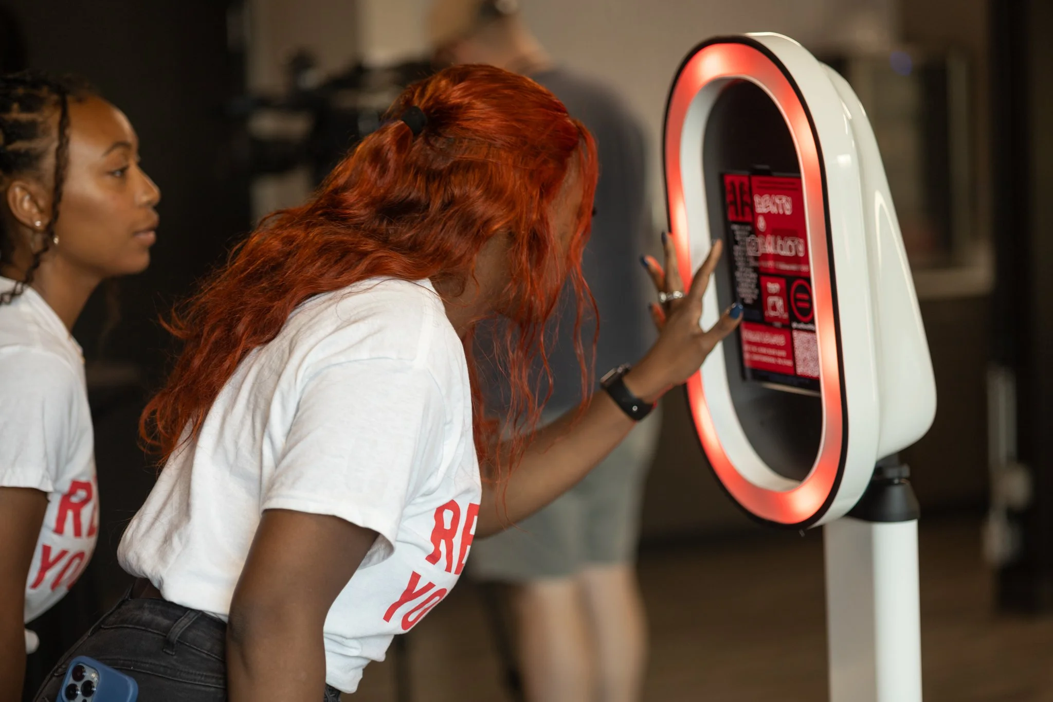 Two women wearing white t-shirts with red text, one with curly hair and the other with long red hair, interact with a tech kiosk with a circular design featuring red lighting.