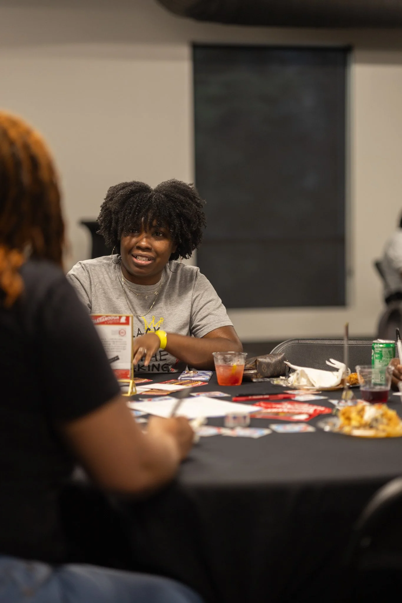A woman with curly hair is engaged in conversation at a table with other people during a gathering. The table has various food and drinks on it.