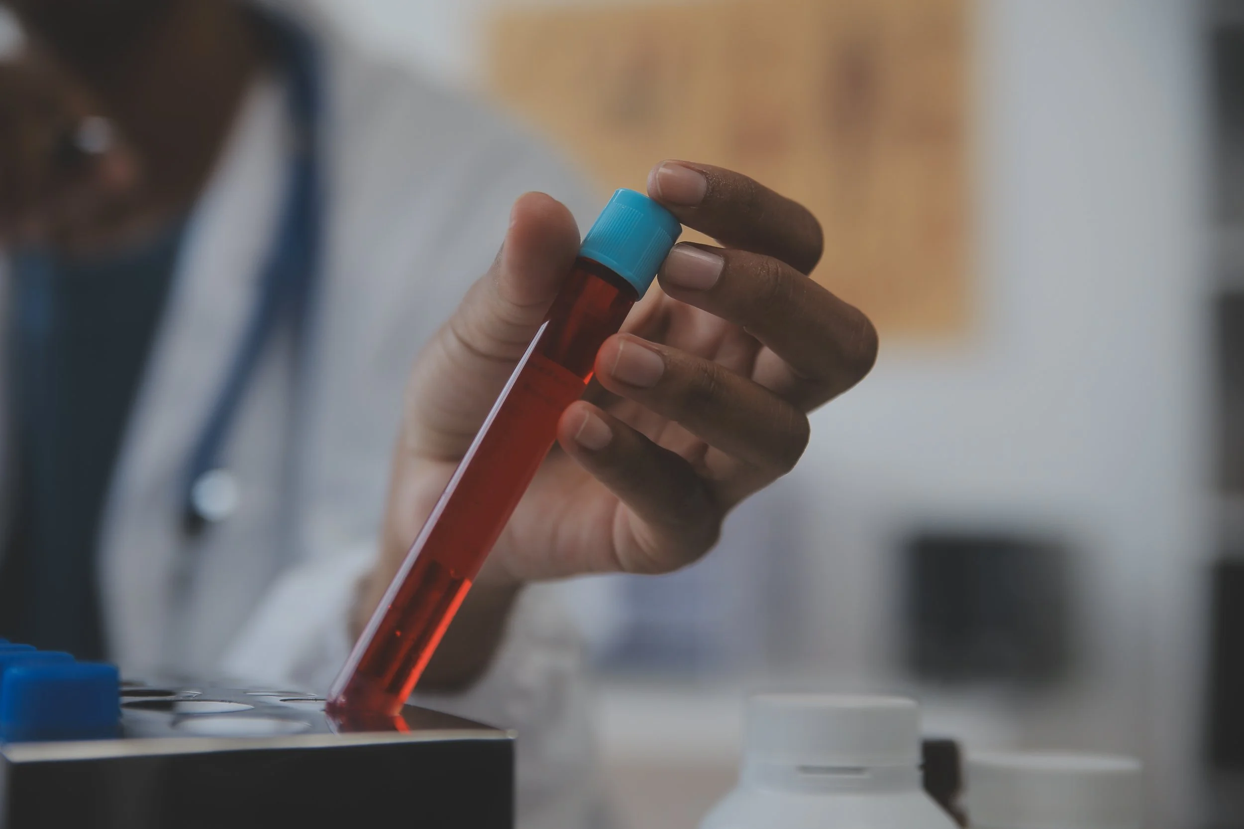 A person wearing a white lab coat holding a test tube filled with red liquid in a laboratory setting.