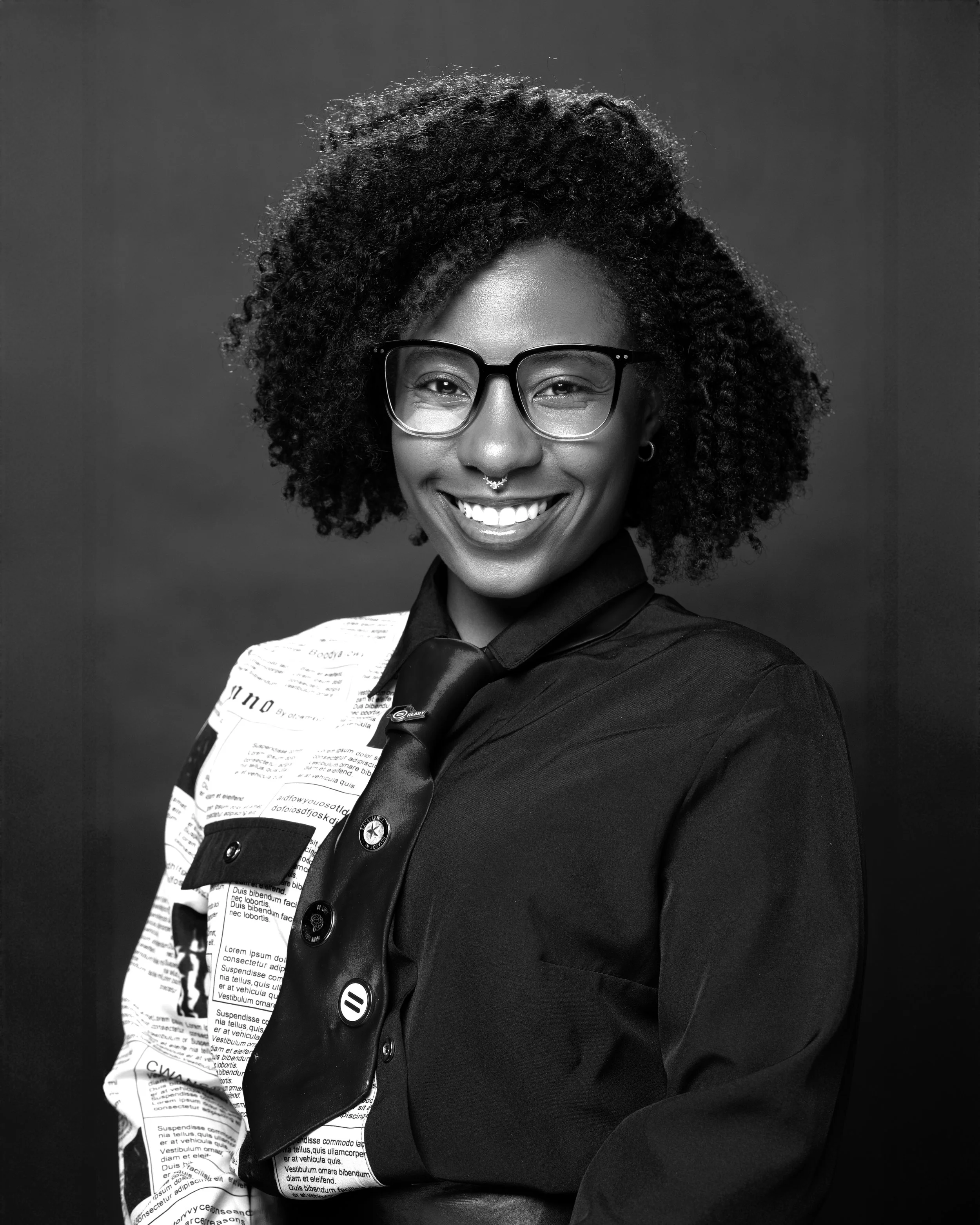 A smiling woman with glasses and curly hair, wearing a black shirt with newspaper print on one side and a black tie, against a plain dark background.