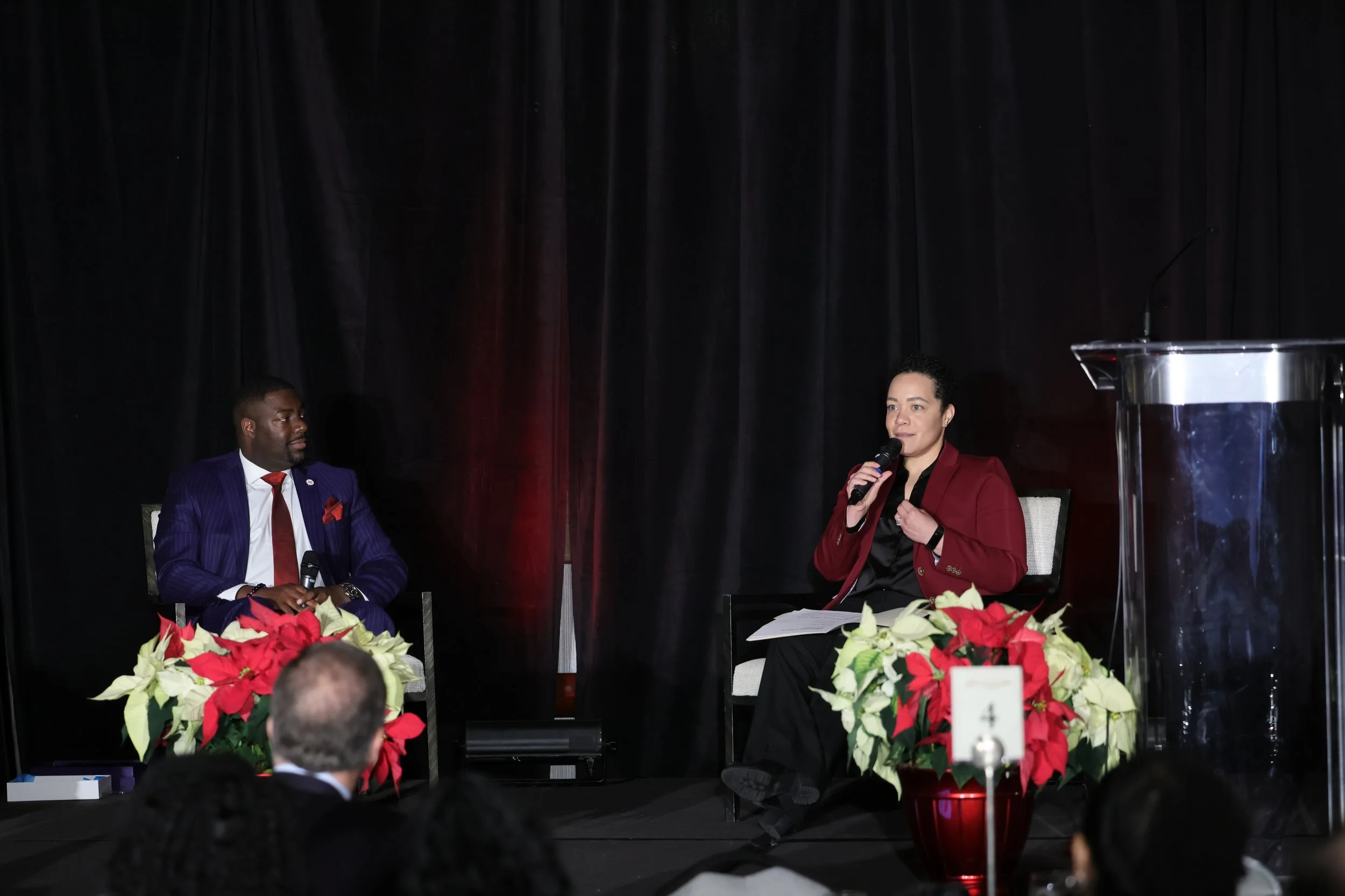 A man and woman seated on stage during a panel discussion, with the woman speaking into a microphone. The stage is decorated with potted poinsettias and a dark curtain backdrop.