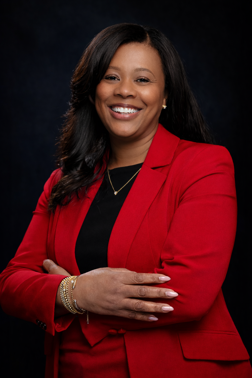 Portrait of a smiling woman with black hair wearing a red blazer, black top, and gold jewelry against a dark background.