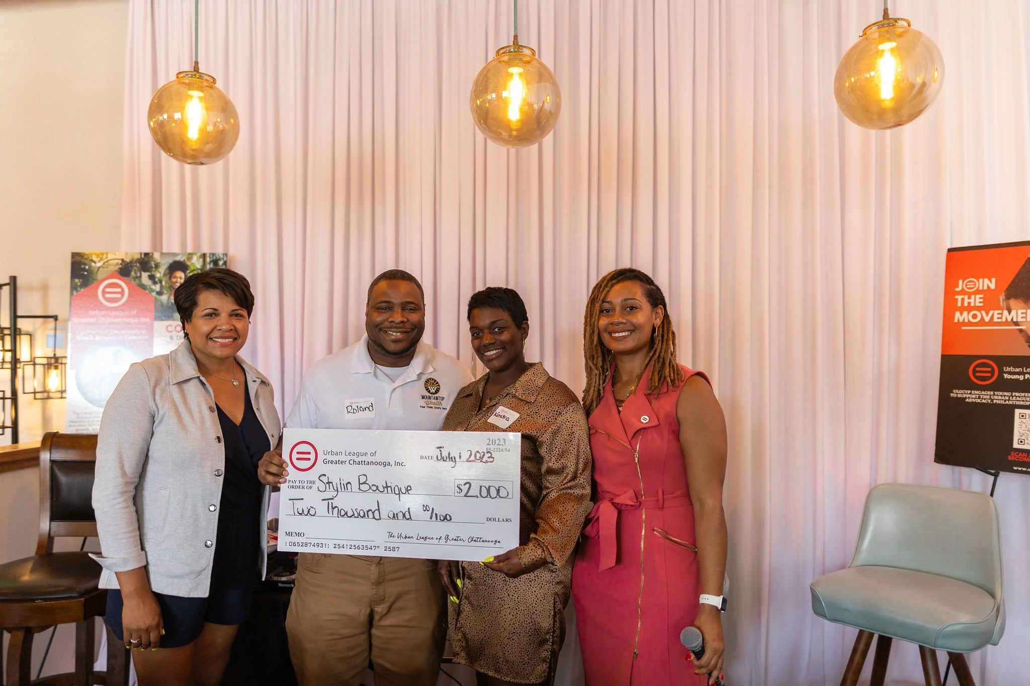 Four women and one man standing together indoors, holding a large check for $2,000 made out to Stylein Boutique during an event. The background features pink curtains and hanging pendant lights.