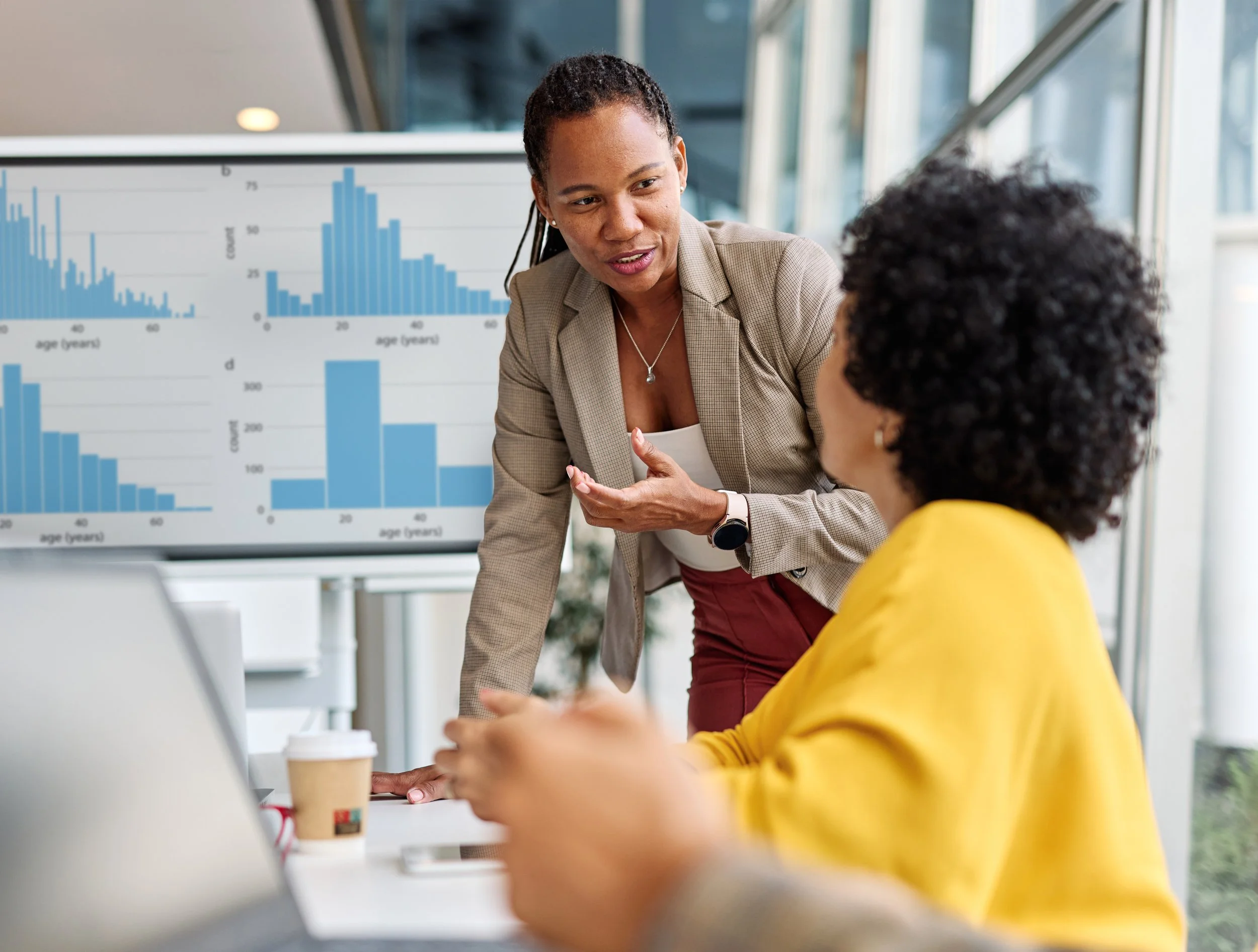 Two women having a business discussion in an office with spreadsheets showing on a screen behind them.