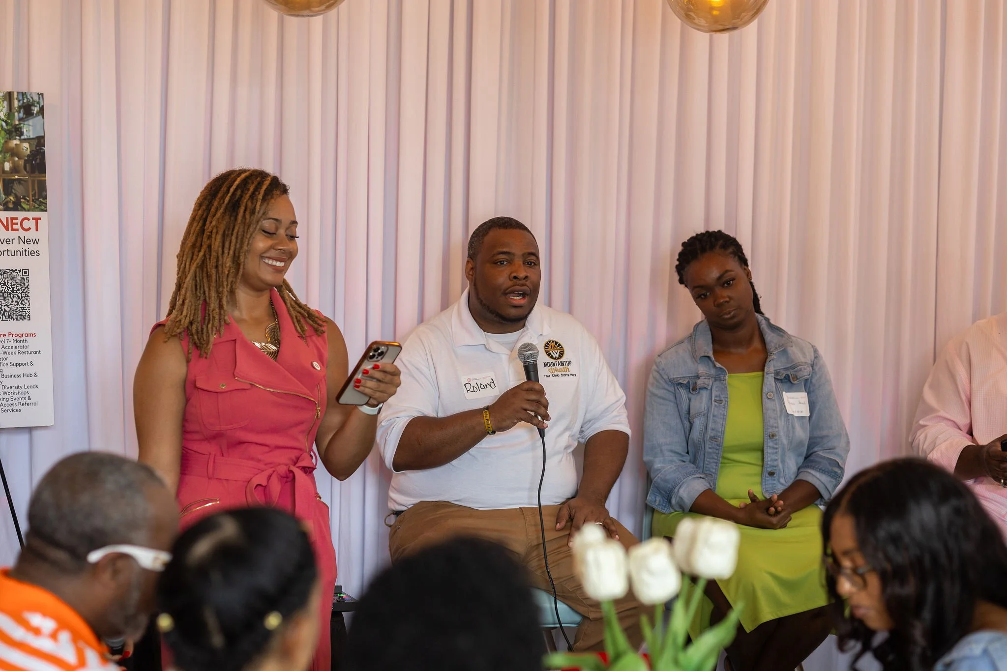 People attending a panel discussion or presentation, with a woman in pink holding a smartphone, a man in front with a microphone, and a woman in a denim jacket listening, set against a white curtain backdrop.