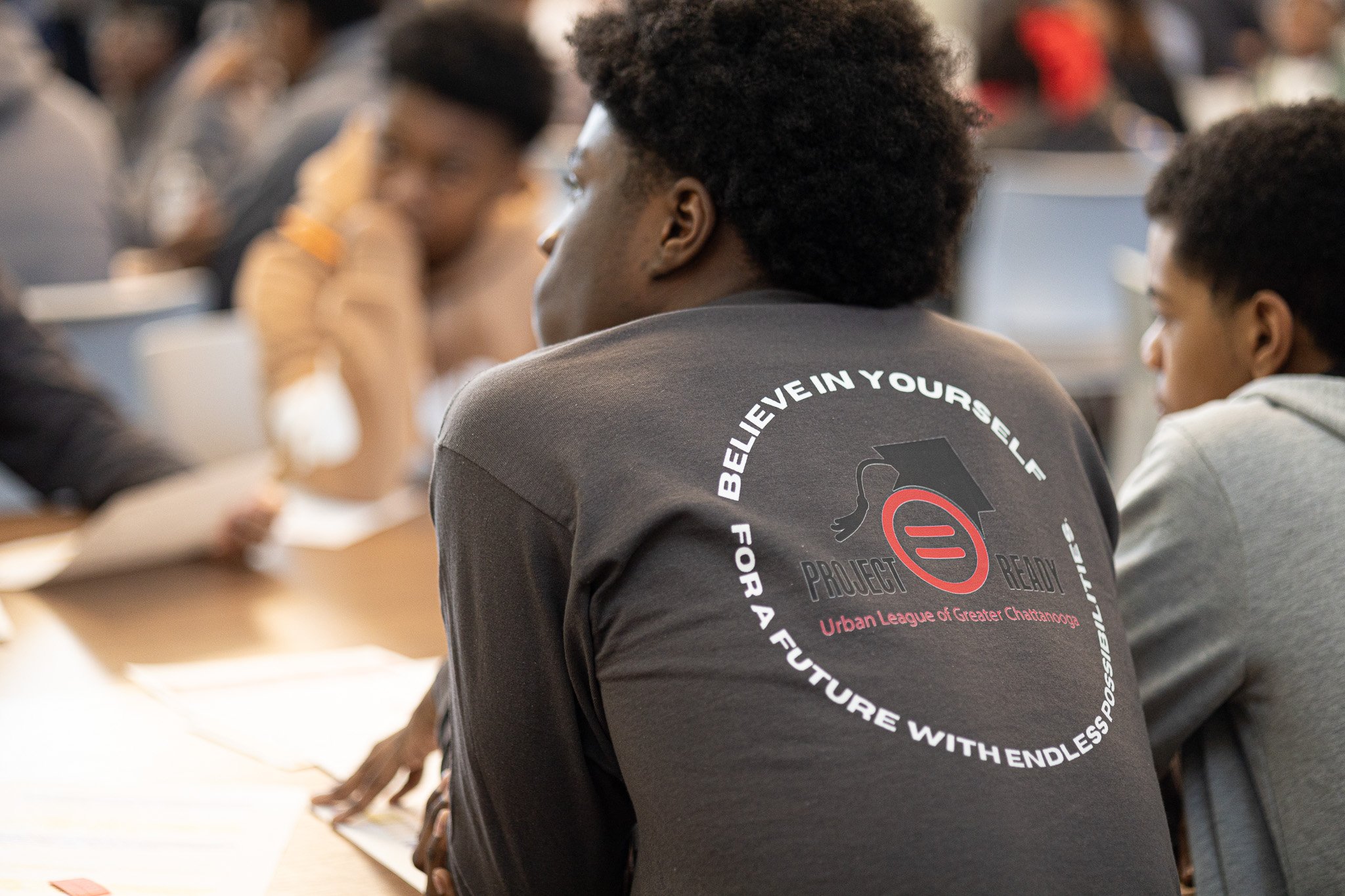 Young man with curly hair wearing a black T-shirt with a logo and circle with the text 'Believe in Yourself for a Future with Endless Possibilities,' sitting at a table with other people in a classroom or conference setting.