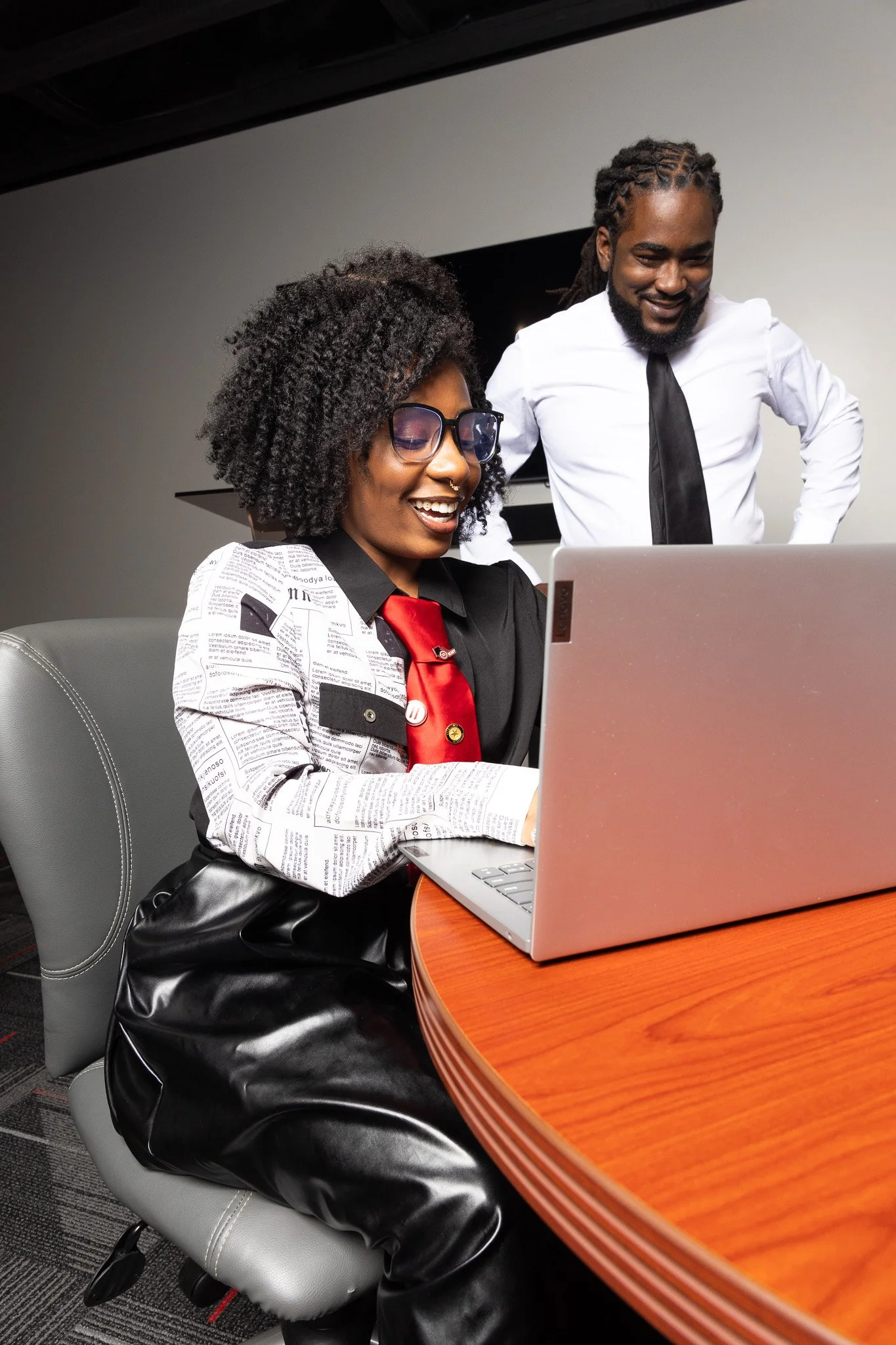 A woman with glasses and curly hair looking at a laptop, smiling, with a man standing beside her, in a business setting.