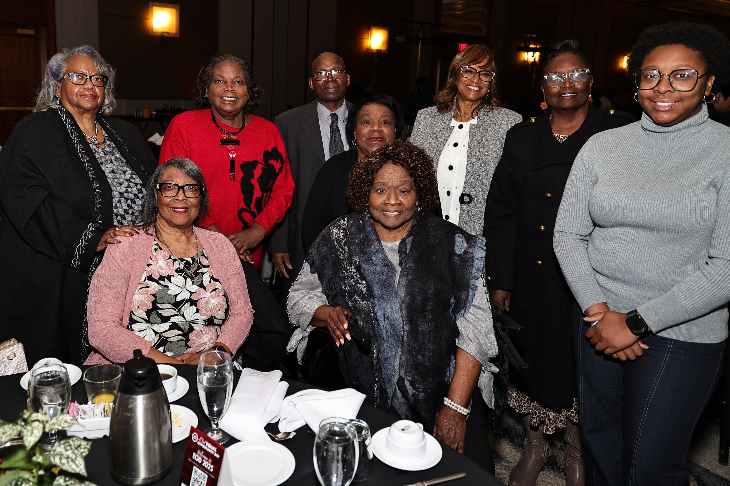 A group of nine women gathered around a table at a formal event. Some women are standing, and one woman is seated in a wheelchair. The table is set with glasses, plates, silverware, and a pitcher, indicating a meal is being served.