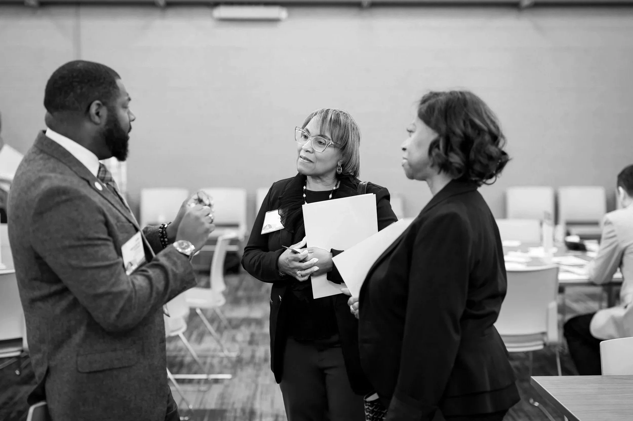 Three professionally dressed people engaged in conversation at a conference or meeting room.