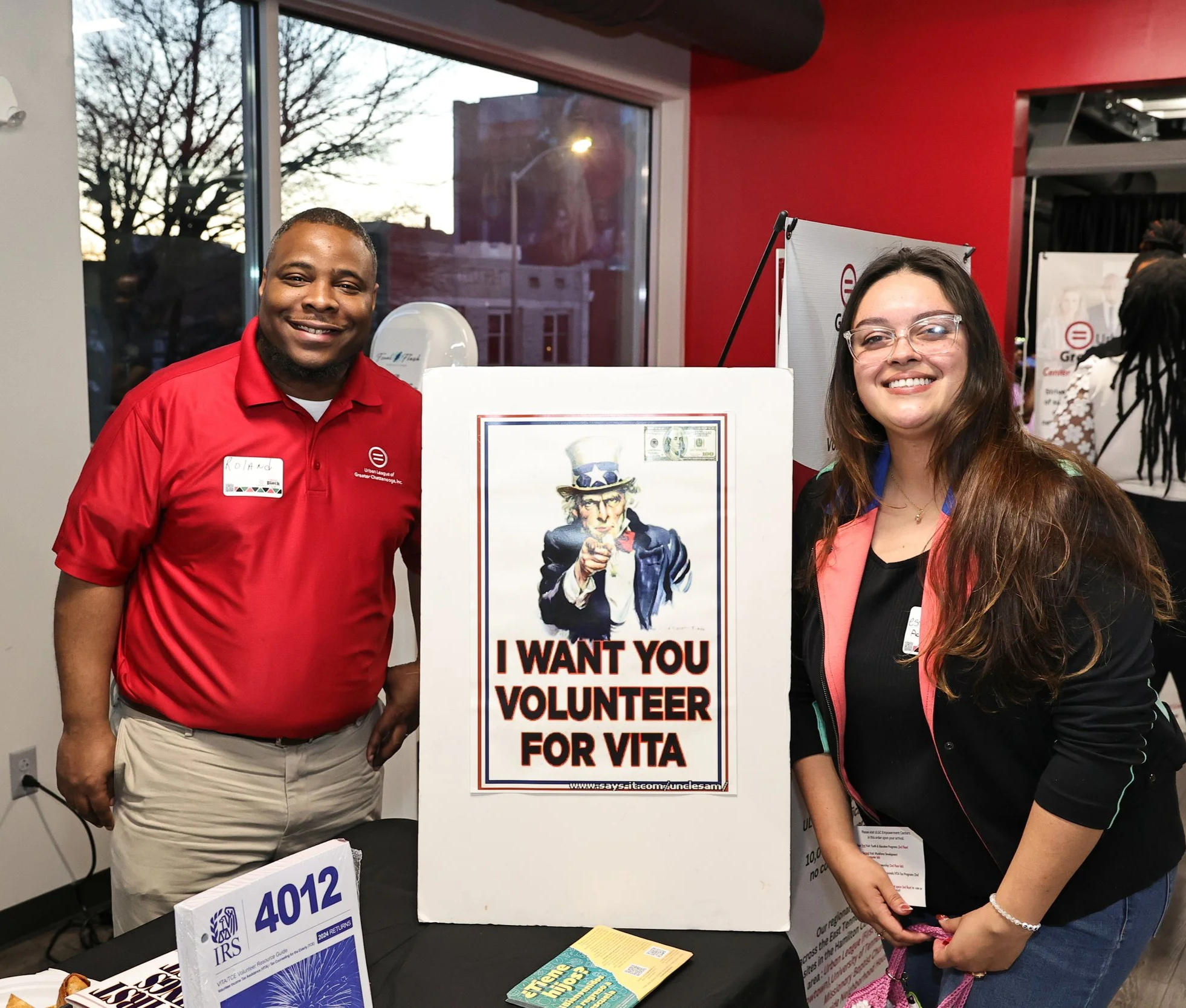 A man and a woman smiling at a volunteer event, standing beside a sign that features Uncle Sam pointing with the text "I WANT YOU VOLUNTEER FOR VITA." The man is wearing a red polo shirt with a name tag, and the woman has long brown hair, glasses, and a black jacket with pink accents. They are indoors, and there is a large window behind them showing a sunset or dusk sky.