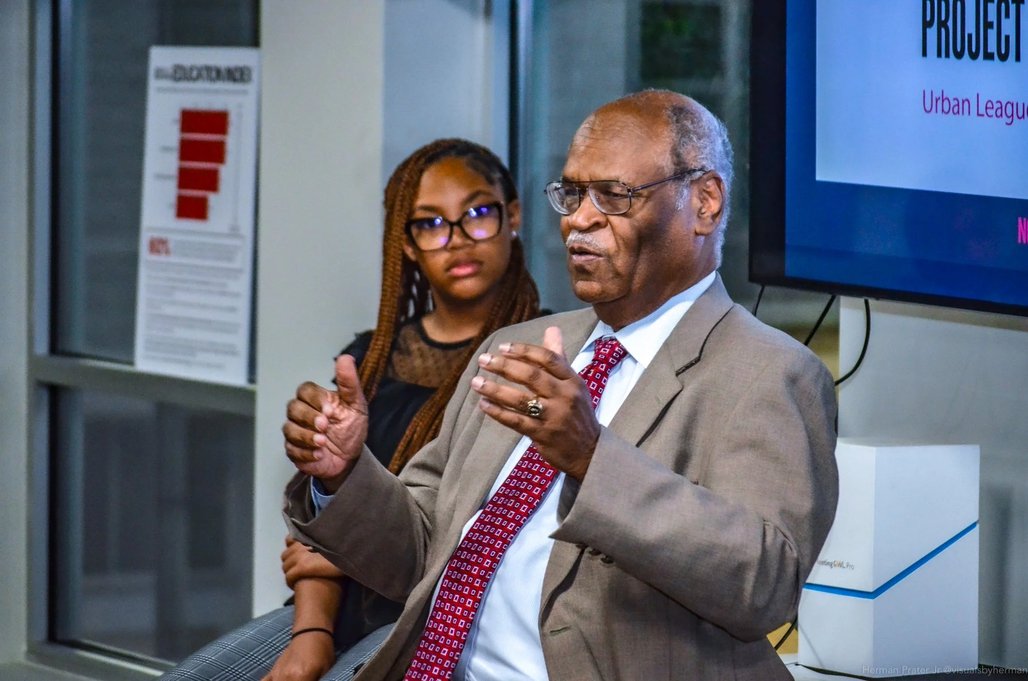 An elderly man in a beige suit and red tie is speaking and gesturing with his hands during a presentation, with a young woman with braided hair and glasses listening attentively behind him. A large screen displays a PowerPoint slide.
