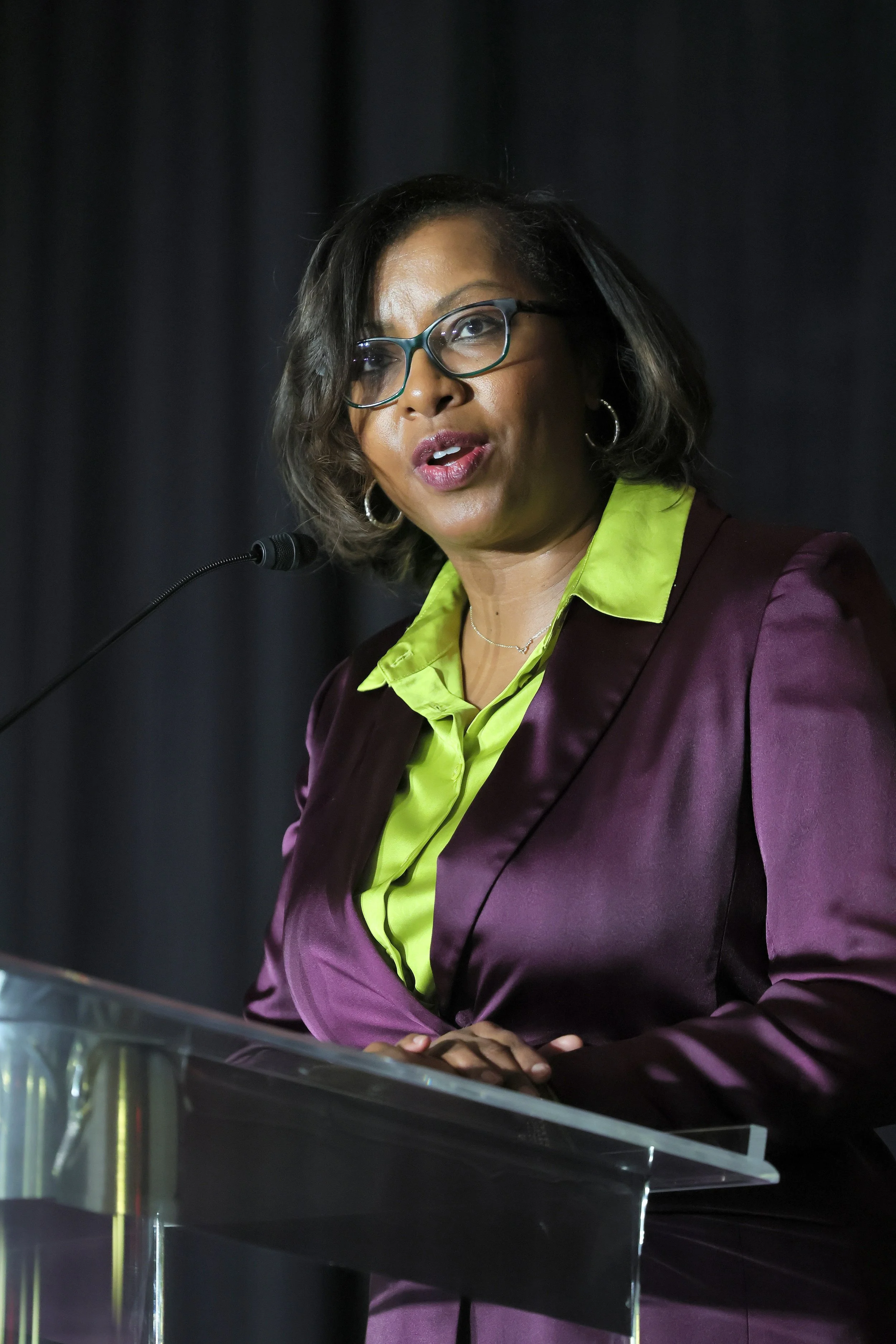 A woman speaking at a podium, wearing glasses, a lime green shirt, and a purple blazer, with dark hair and earrings.