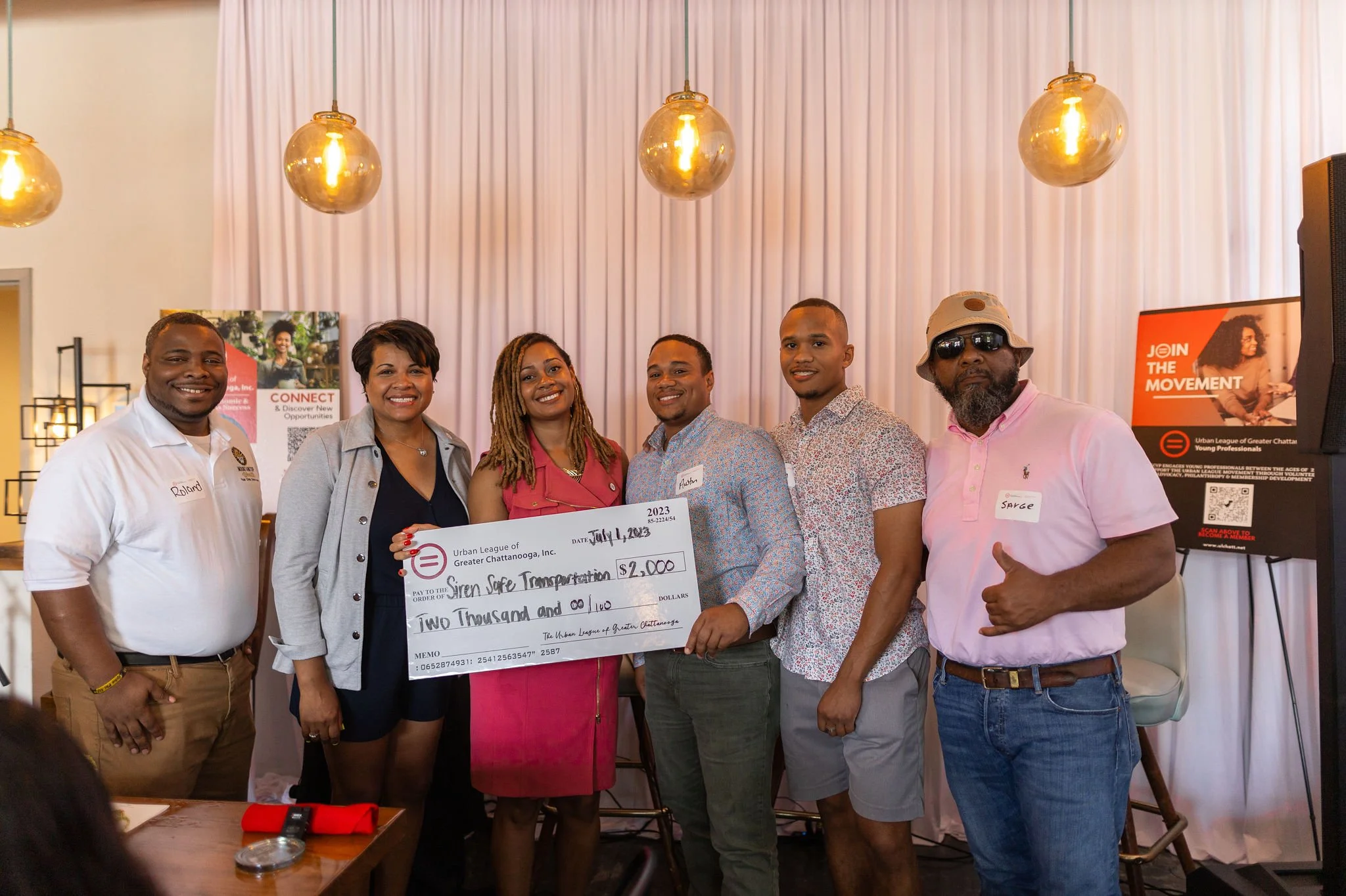 Group of seven people standing together at an event, holding a large check for $2,000 made out to Siren Safe Transportation, in a room with pink curtains and warm lighting.