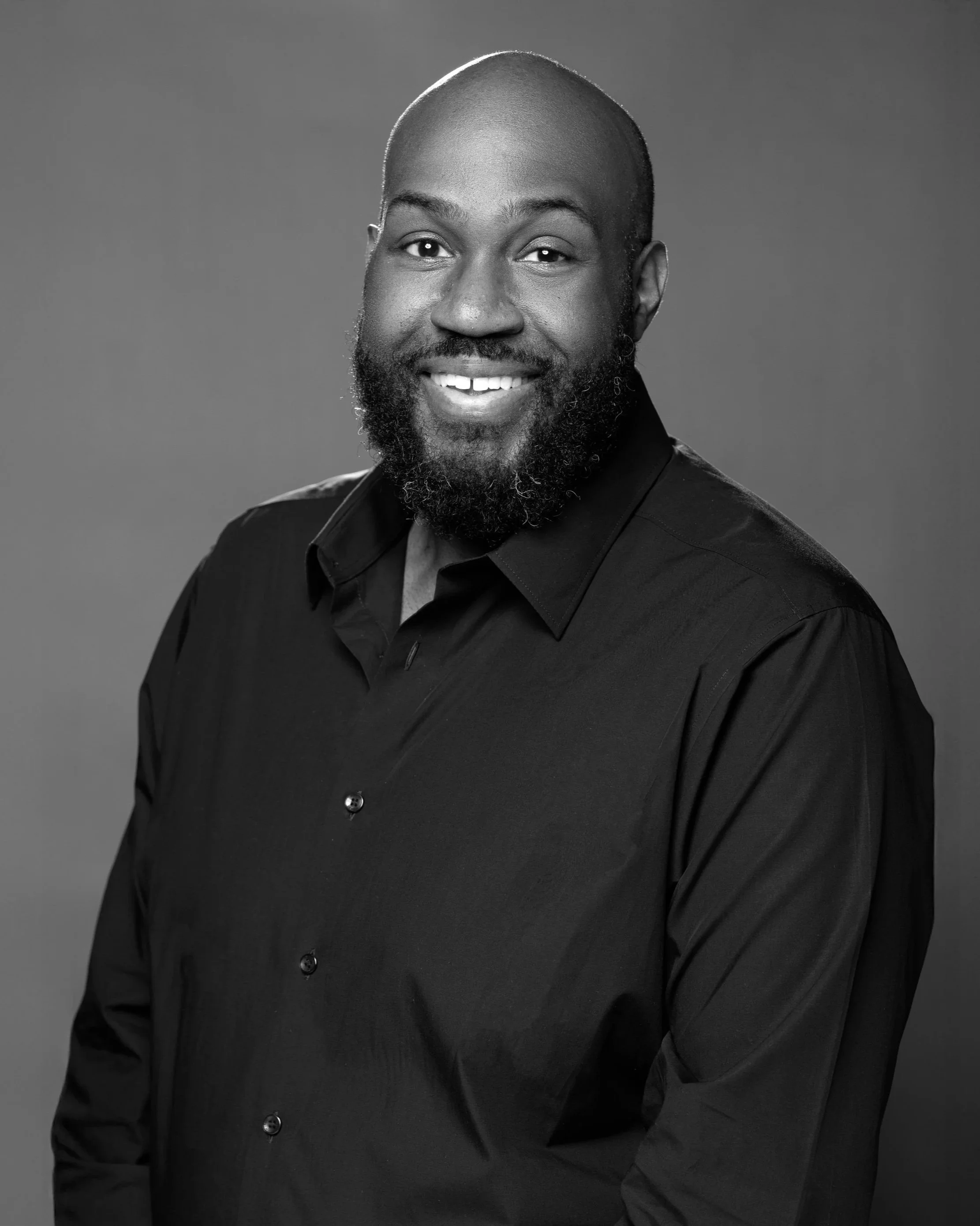 Black-and-white studio portrait of a smiling man with a beard, wearing a black collared shirt, against a plain background.