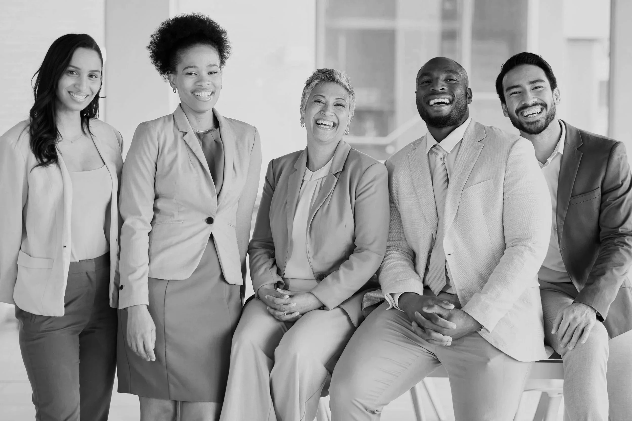 Five diverse professional individuals smiling and posing together in an office setting, with two seated and three standing.