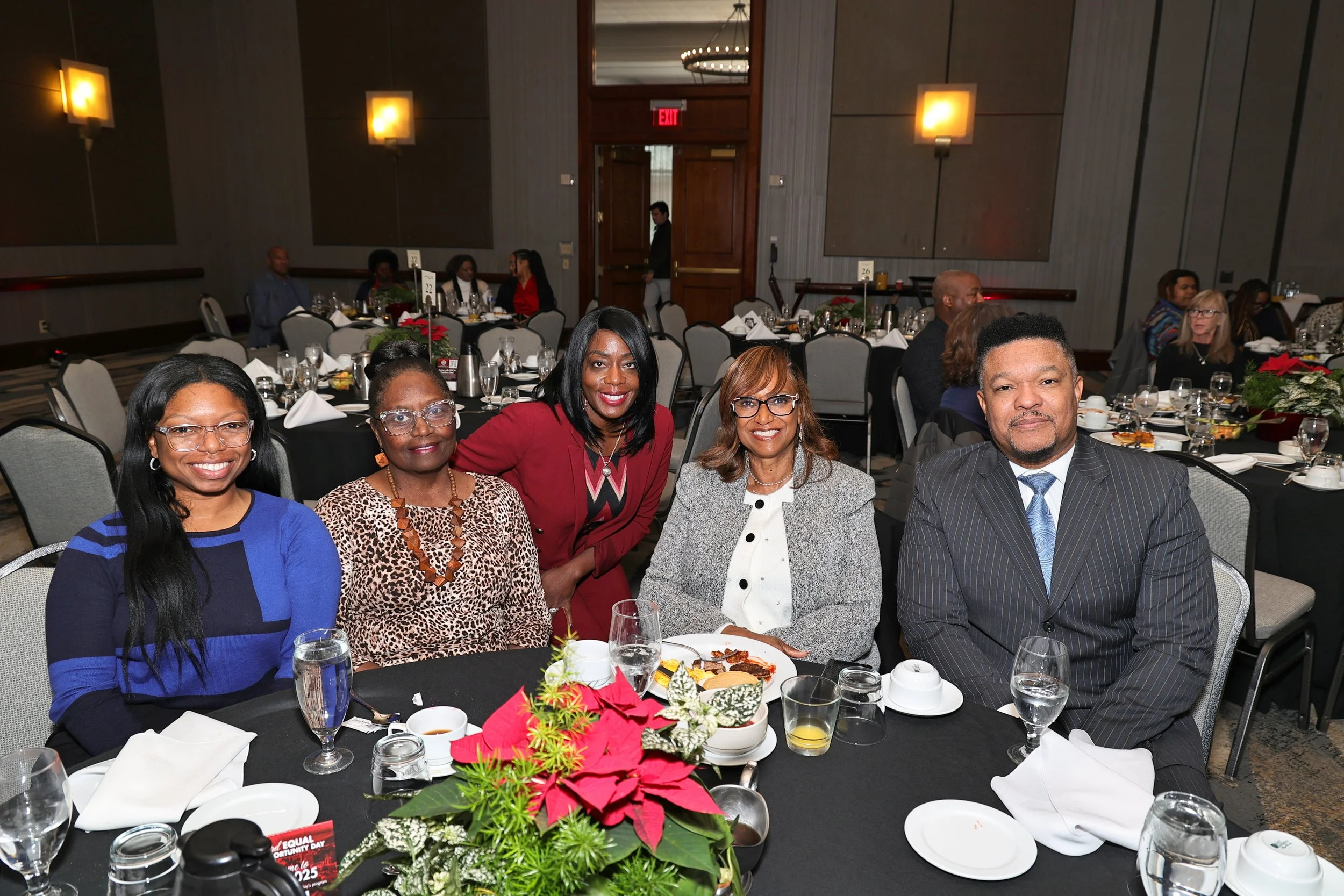 Group of five people, three women and two men, sitting at a round table during a formal event or celebration, with holiday decorations and tables set with food and drinks in the background.