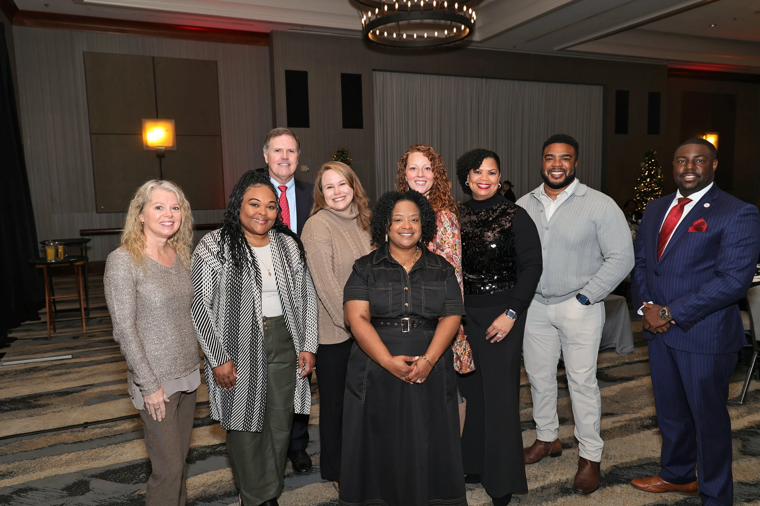 Group of ten diverse professionals smiling in a conference room, some wearing business casual attire, with a decorated Christmas tree in the background.