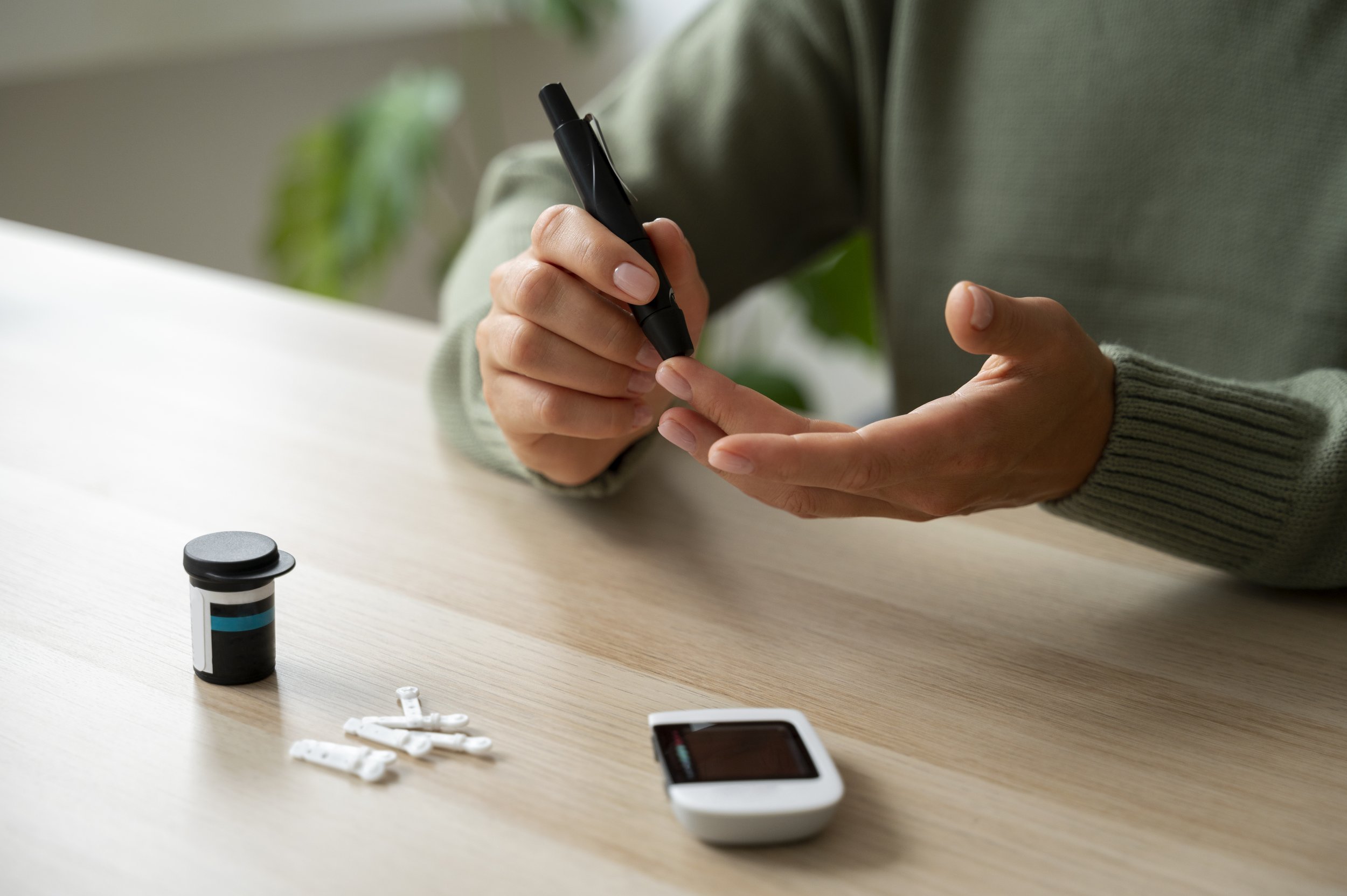 Person testing blood sugar with a glucometer, with a medication bottle and insulin pens on a light wooden table.