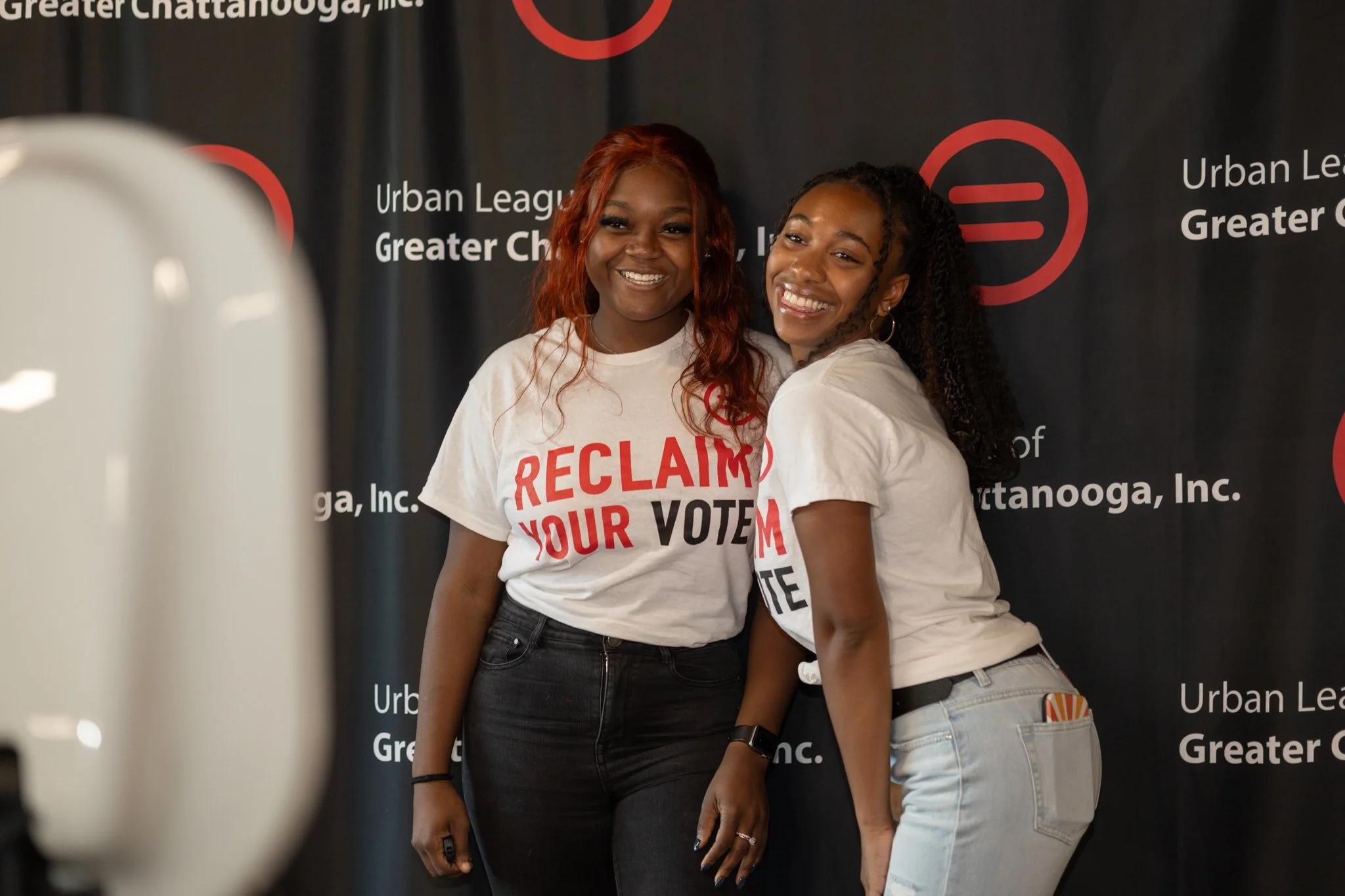 Two women smiling and posing for a photo in front of a black backdrop with the text 'Urban League Greater Chattanooga, Inc.' and a red logo. They are wearing white T-shirts with red and black text that says 'RECLAIM YOUR VOTE'.