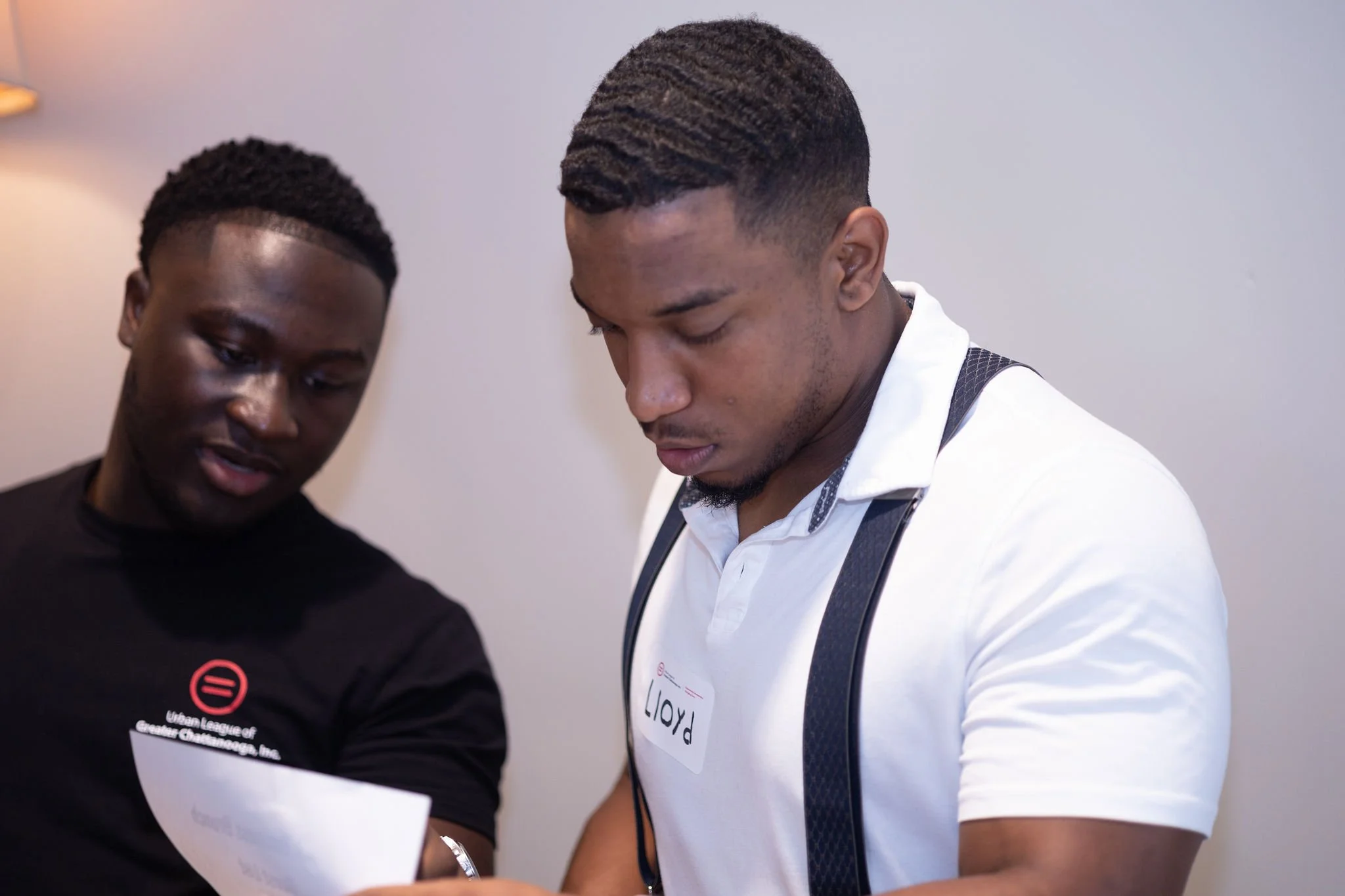 Two young men looking at a piece of paper together, one wearing a black shirt with a logo and the other in a white polo with a name tag that says Lloyd.