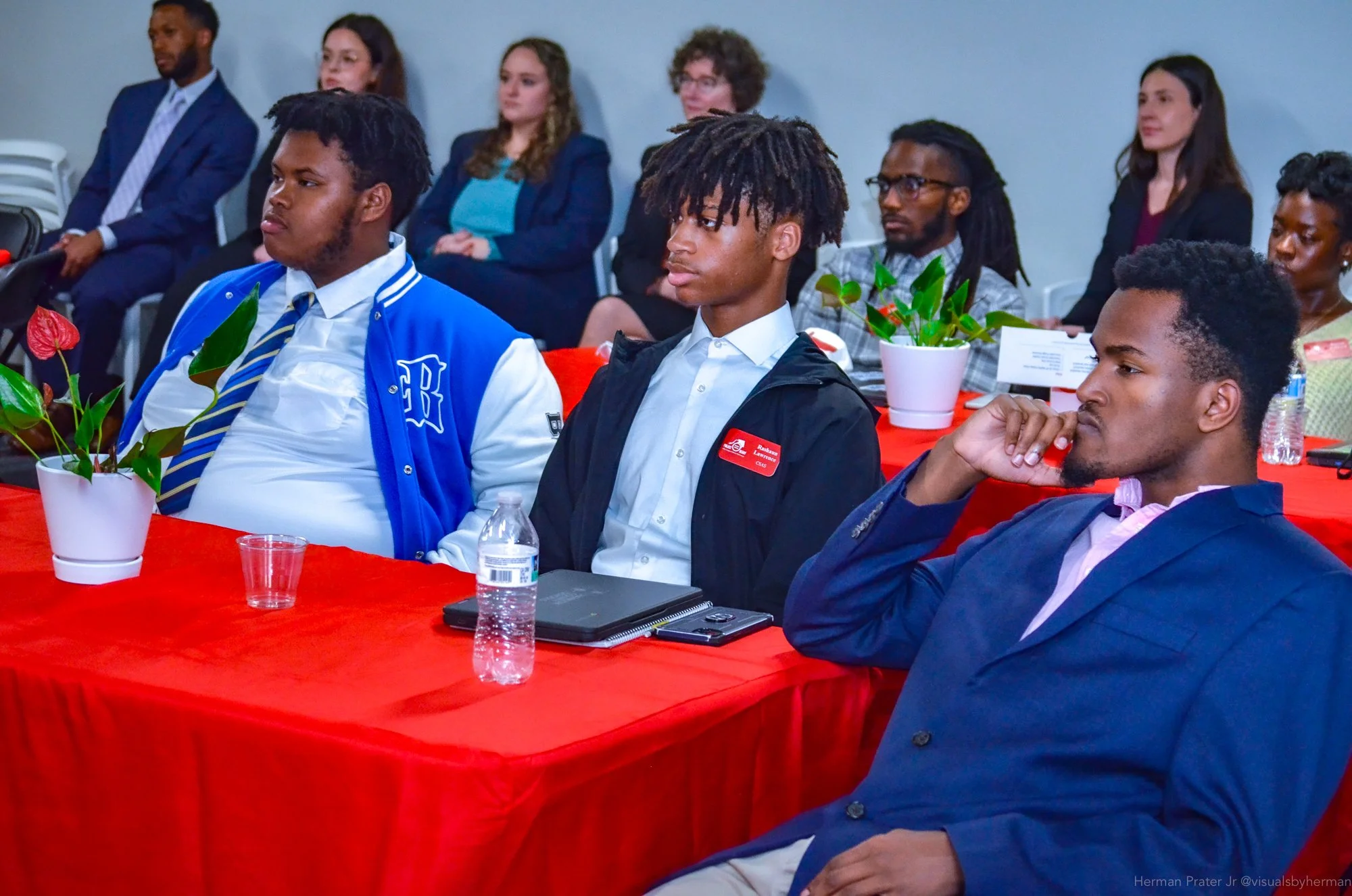 People attending a conference or seminar, sitting at a red-covered table with potted plants, listening attentively to a speaker.