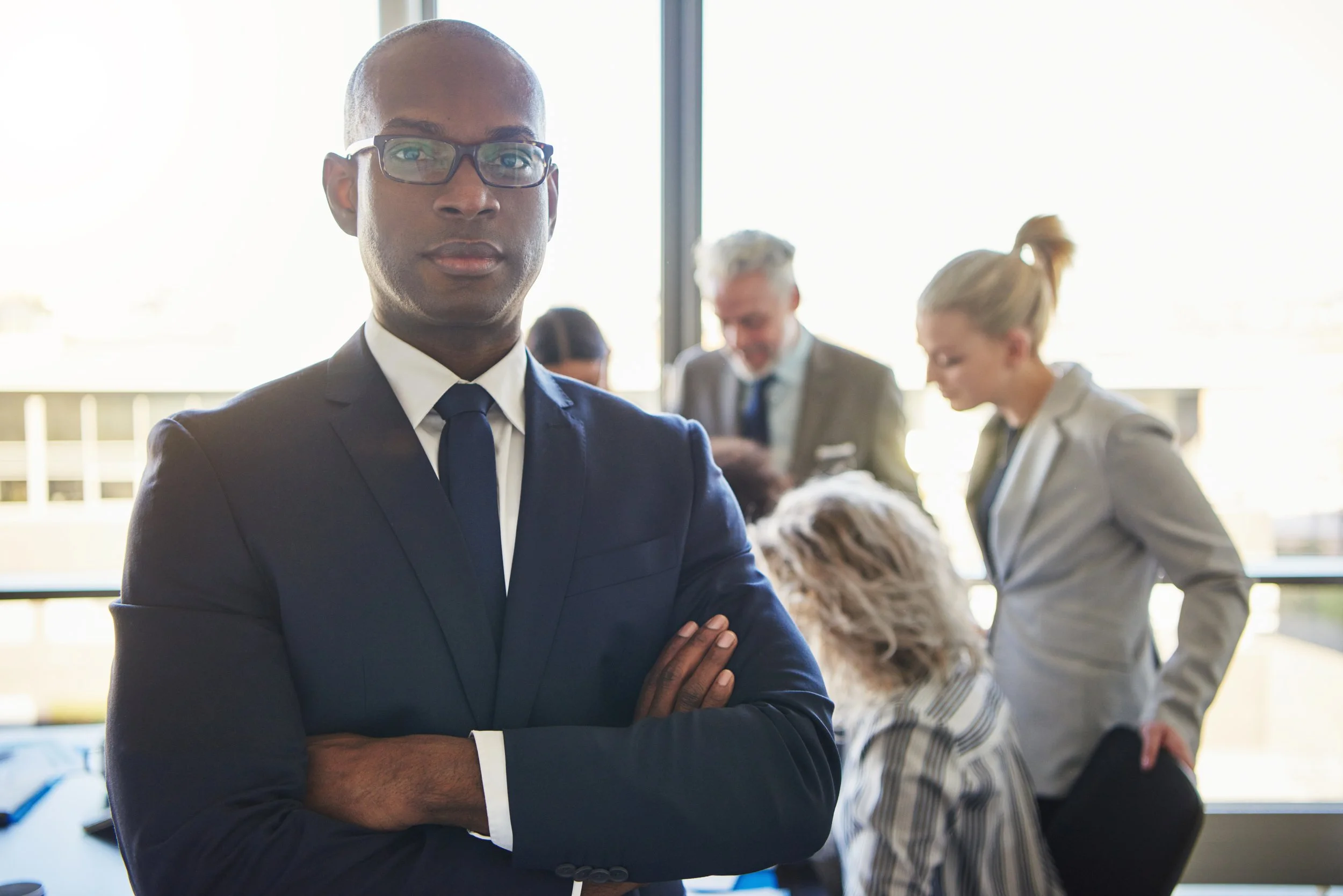 A man in a dark suit with glasses, standing with arms crossed in a business meeting room with other professionals in the background.