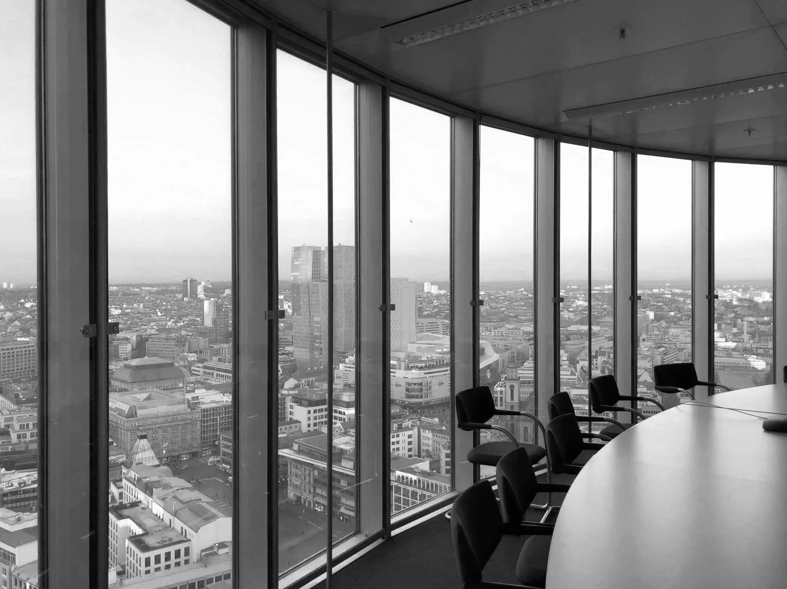 Empty conference room with a large oval table and chairs, overlooking a cityscape through floor-to-ceiling windows.