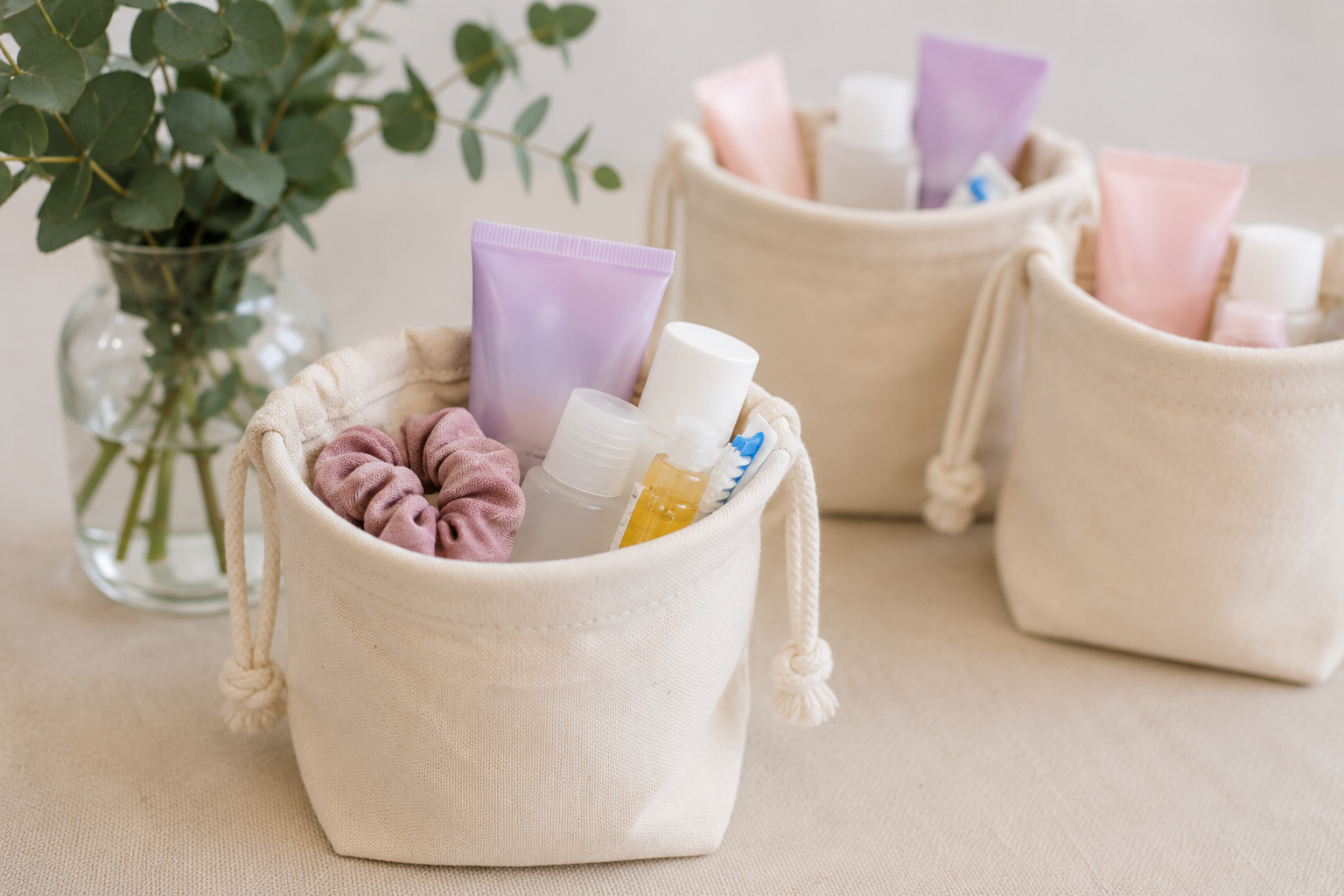 Baskets filled with toiletries such as tubes, bottles, and a toothbrush, placed on a table near a glass vase with green eucalyptus leaves.