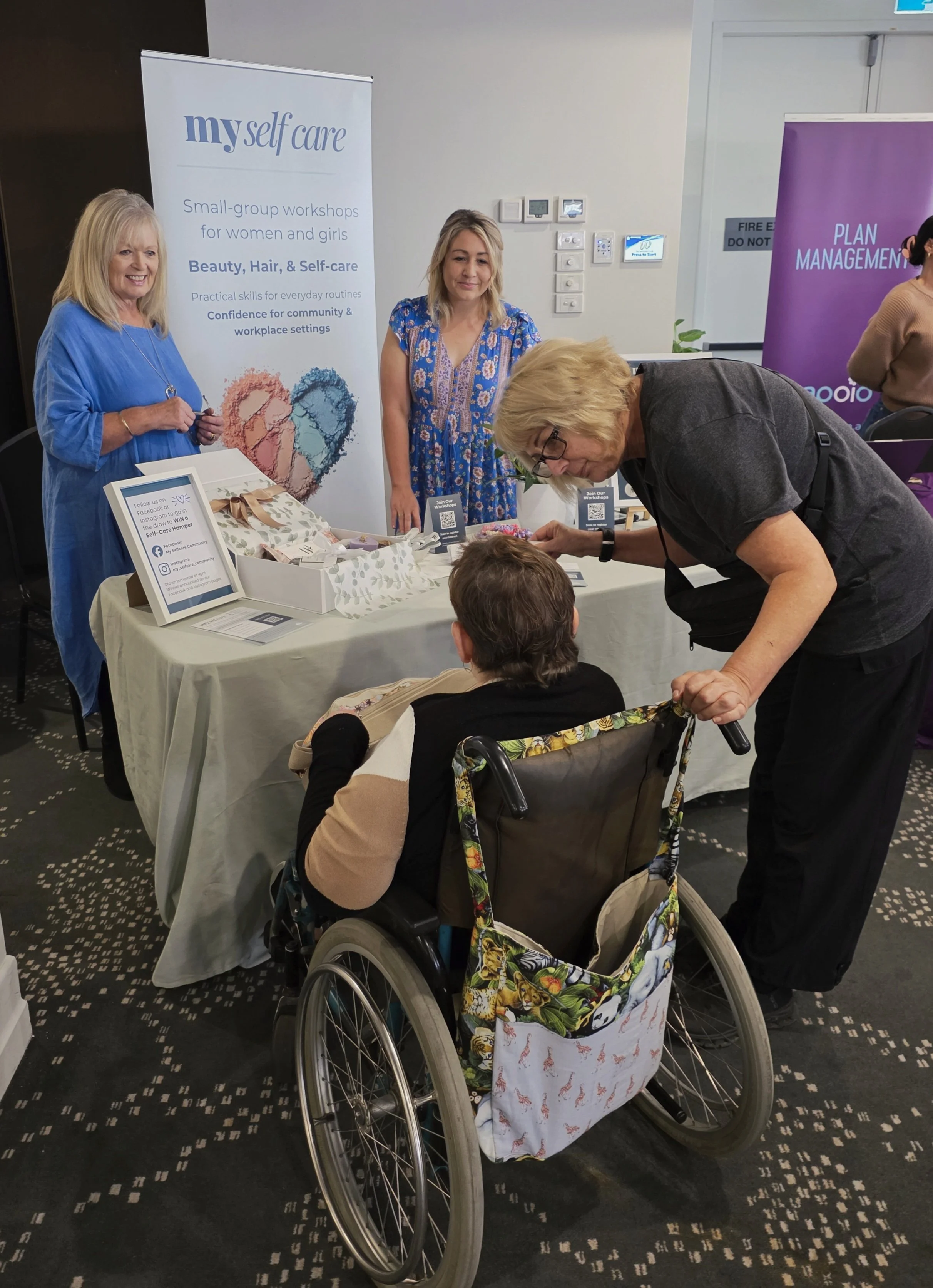 My Selfcare staff attending at an event promoting self-care, beauty, hair, and wellness for women and girls, with informational banners, a person in a wheelchair and their carer.