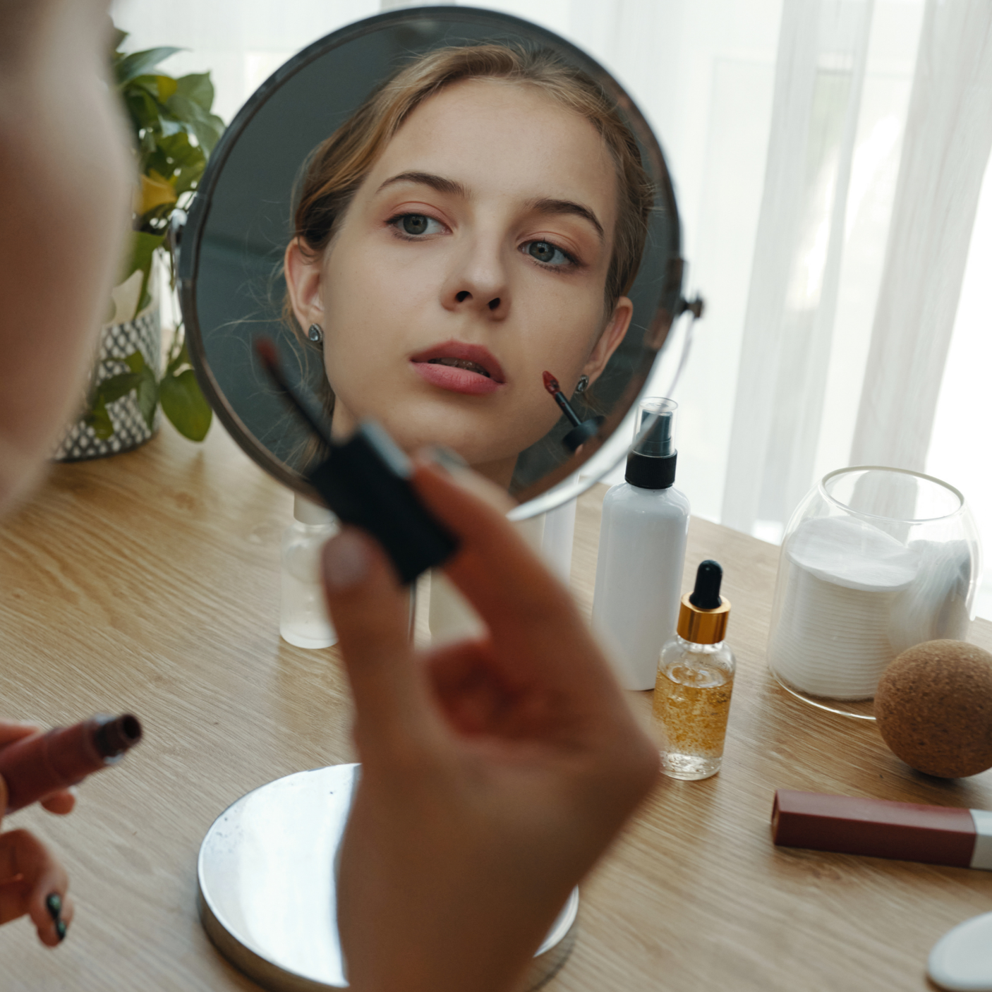 A woman applying lipstick while looking into a mirror on a table with various skincare and makeup products.